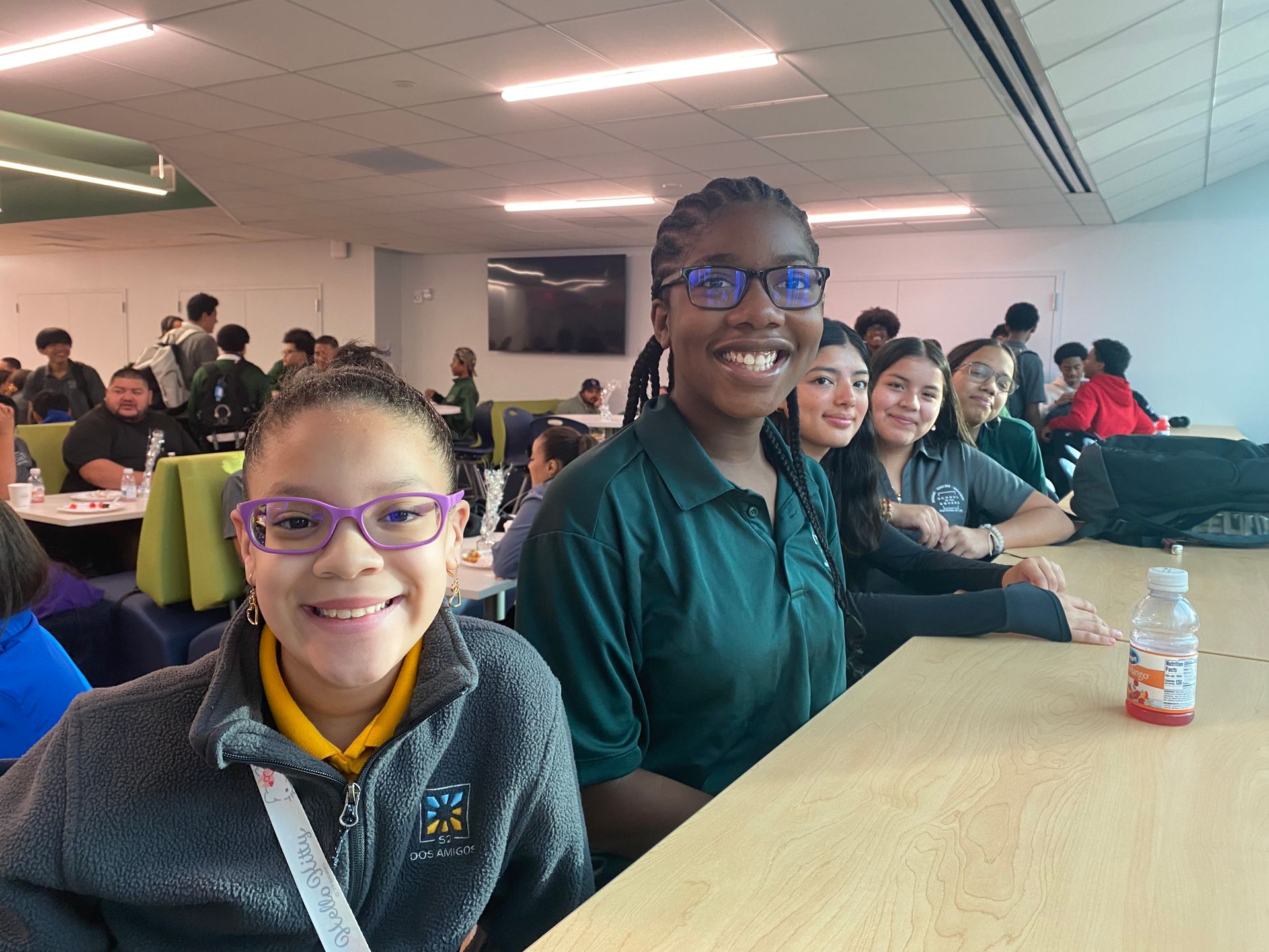 Two smiling students at a long table in a cafeteria, with others seated in the background.