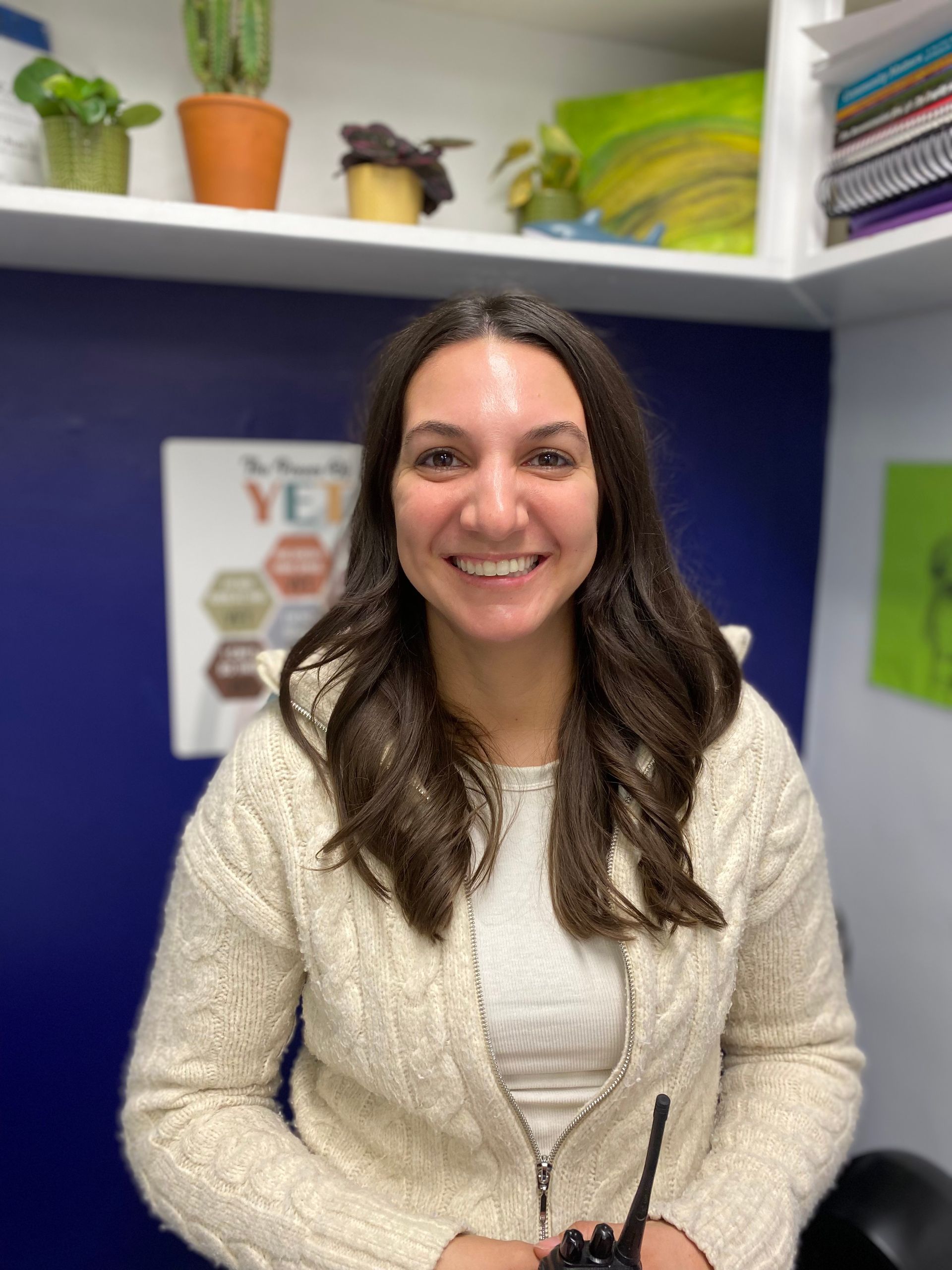 Woman smiling at a desk in an office, with shelves, plants, and a purple wall behind her.