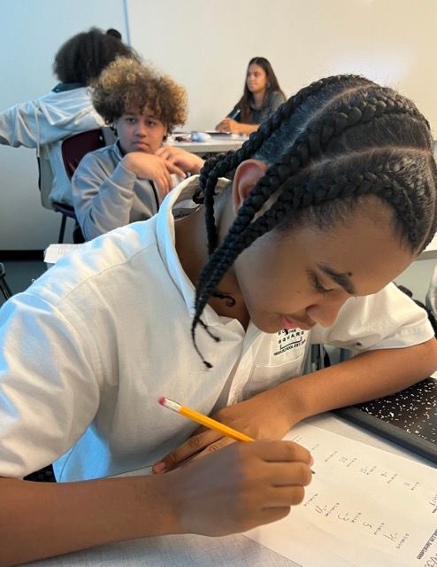 Student writing at a desk in a classroom while classmates work in the background.