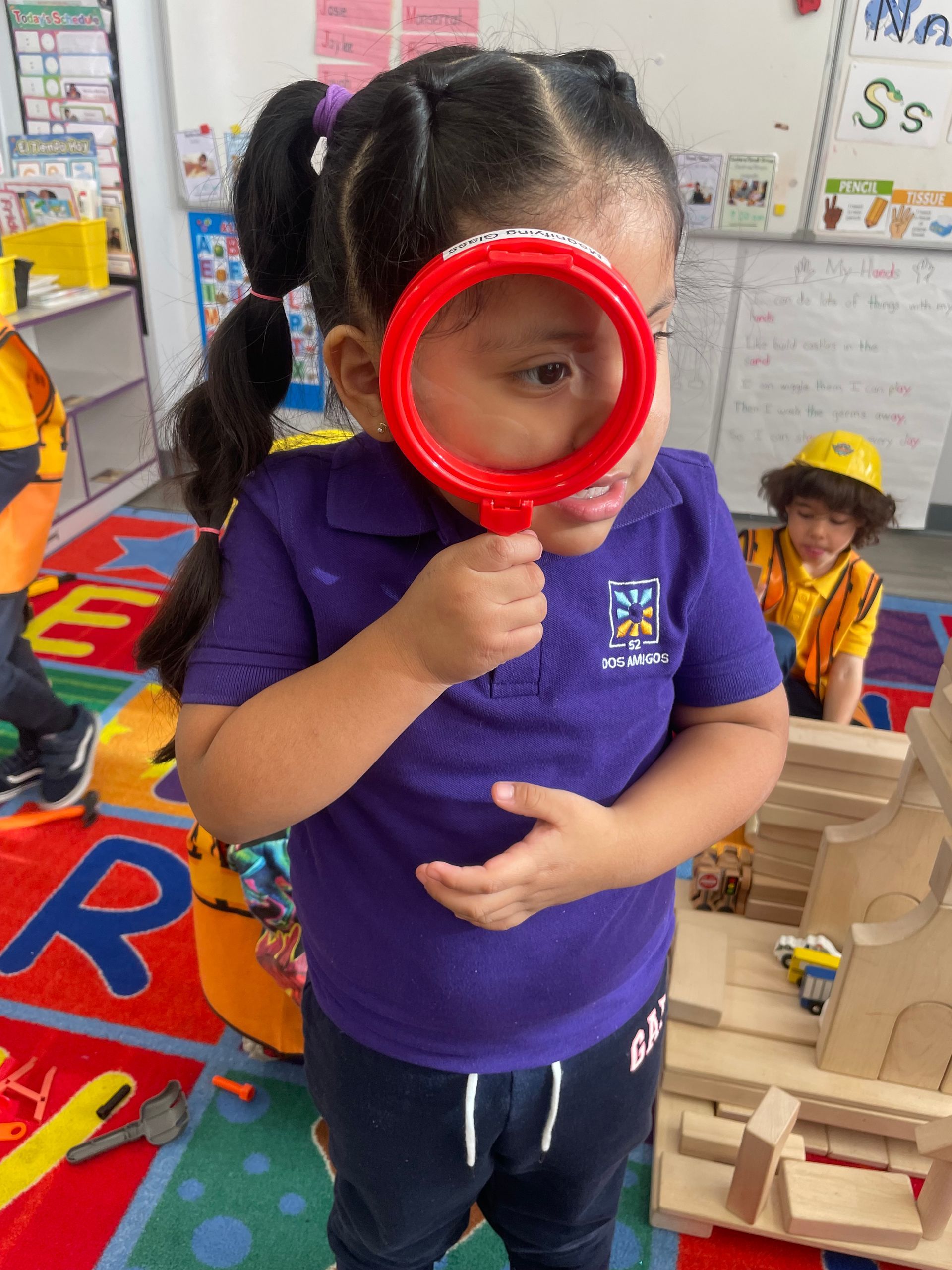Child in purple shirt holding a red magnifying glass in a classroom play area.