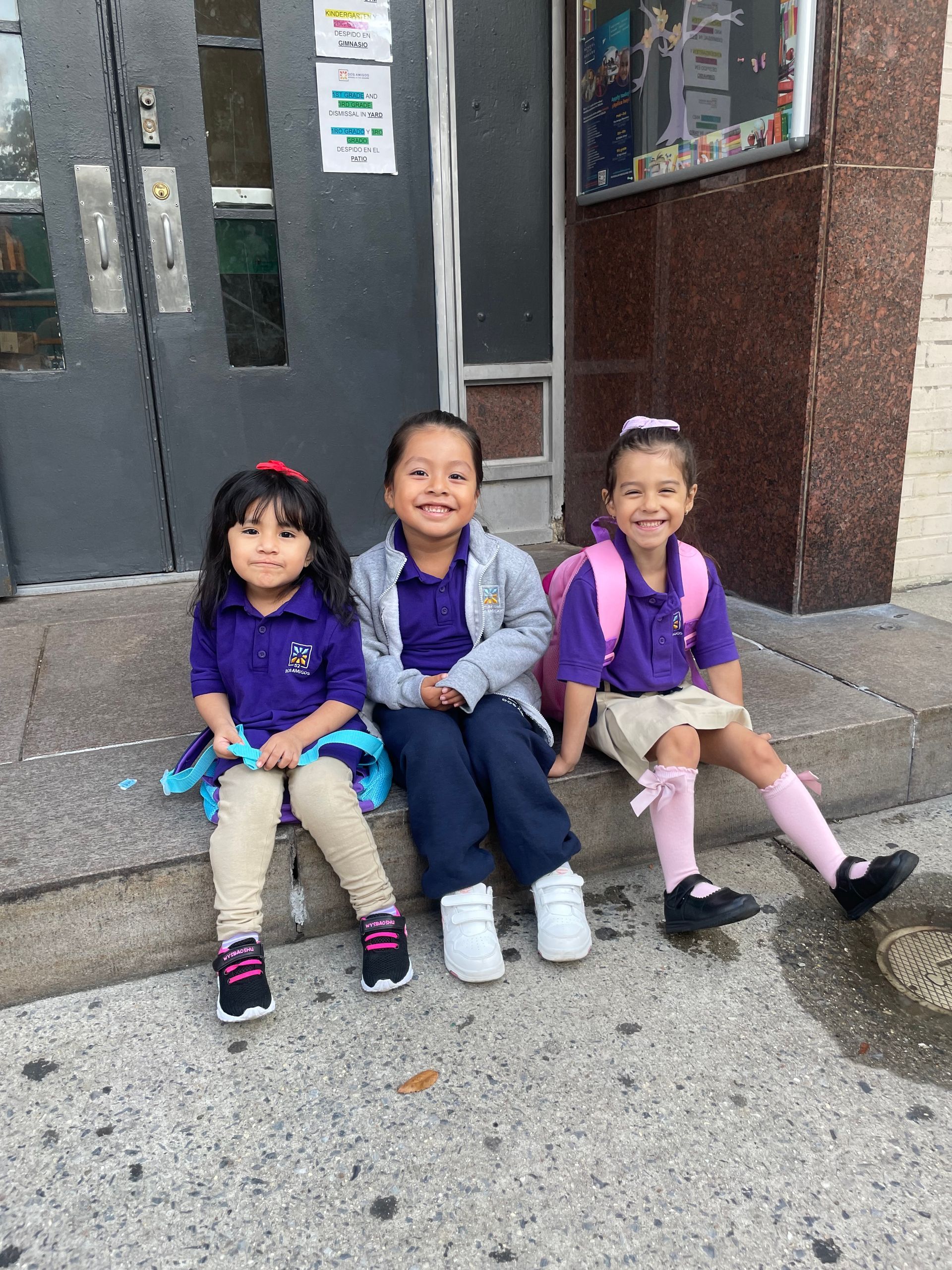 Three children sitting on a sidewalk step outside a building, smiling and wearing colorful jackets.