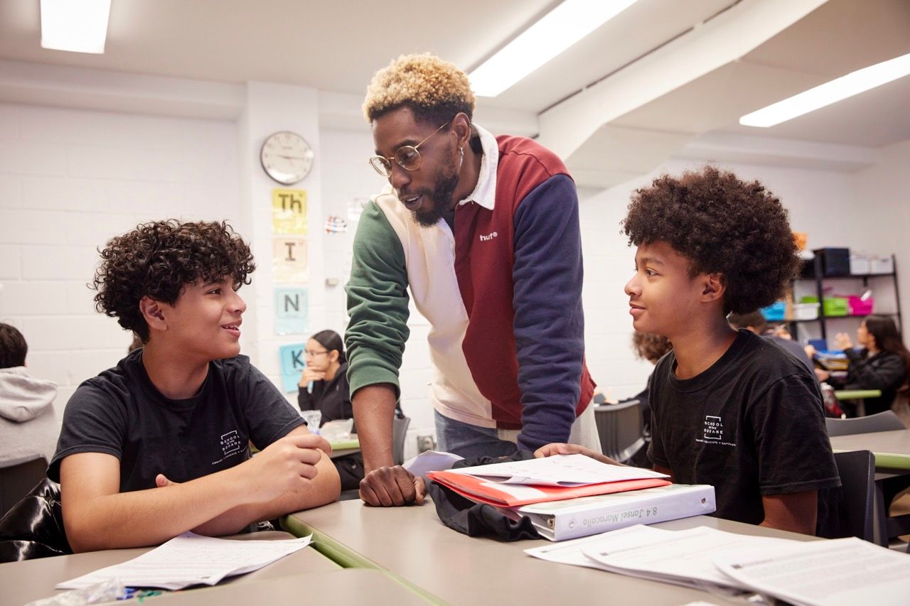 Teacher helping two students at a classroom table with books and papers