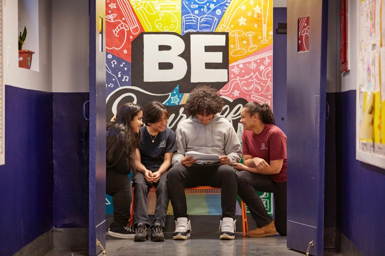 Three people sit on a bench in a hallway beneath a colorful mural with large “BE” text.