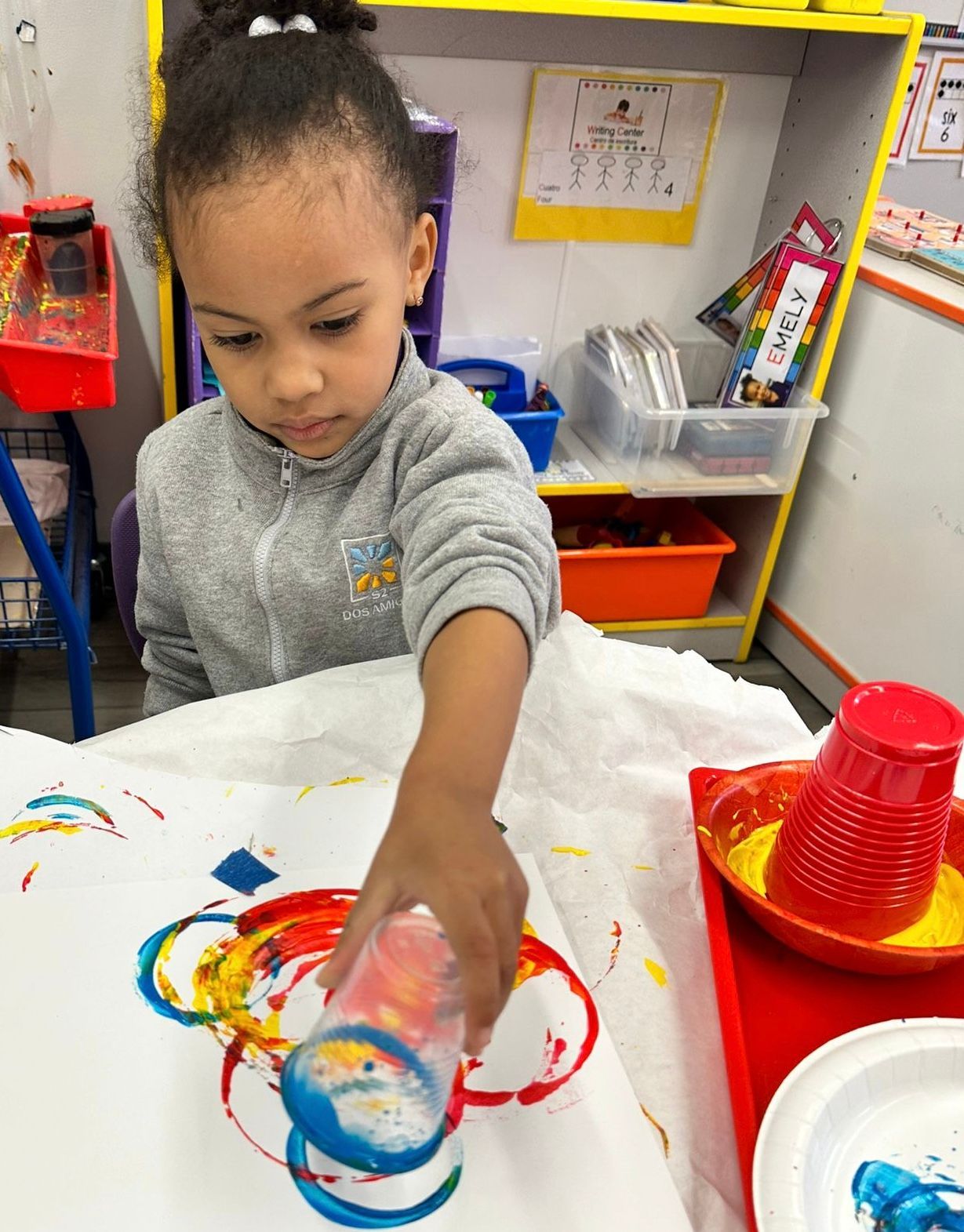 Child painting with colorful handprints at a classroom table with red cups and art supplies.