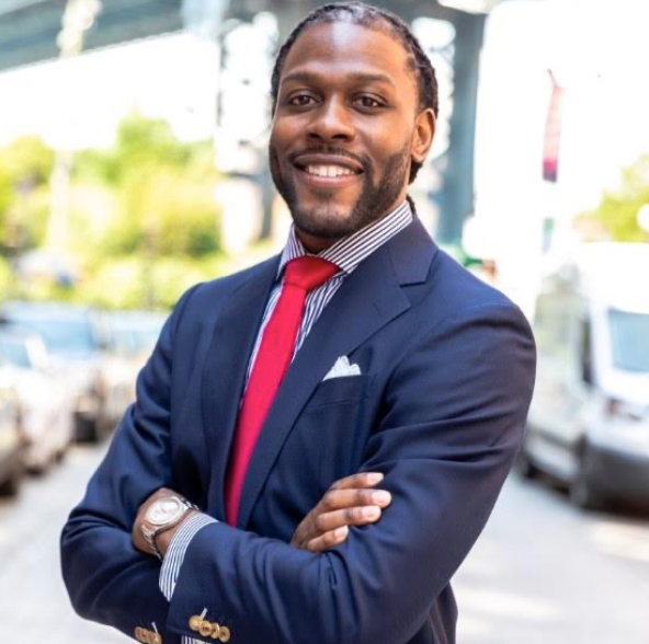 Smiling professional in a navy suit with red tie, arms crossed, standing outdoors on a city street.