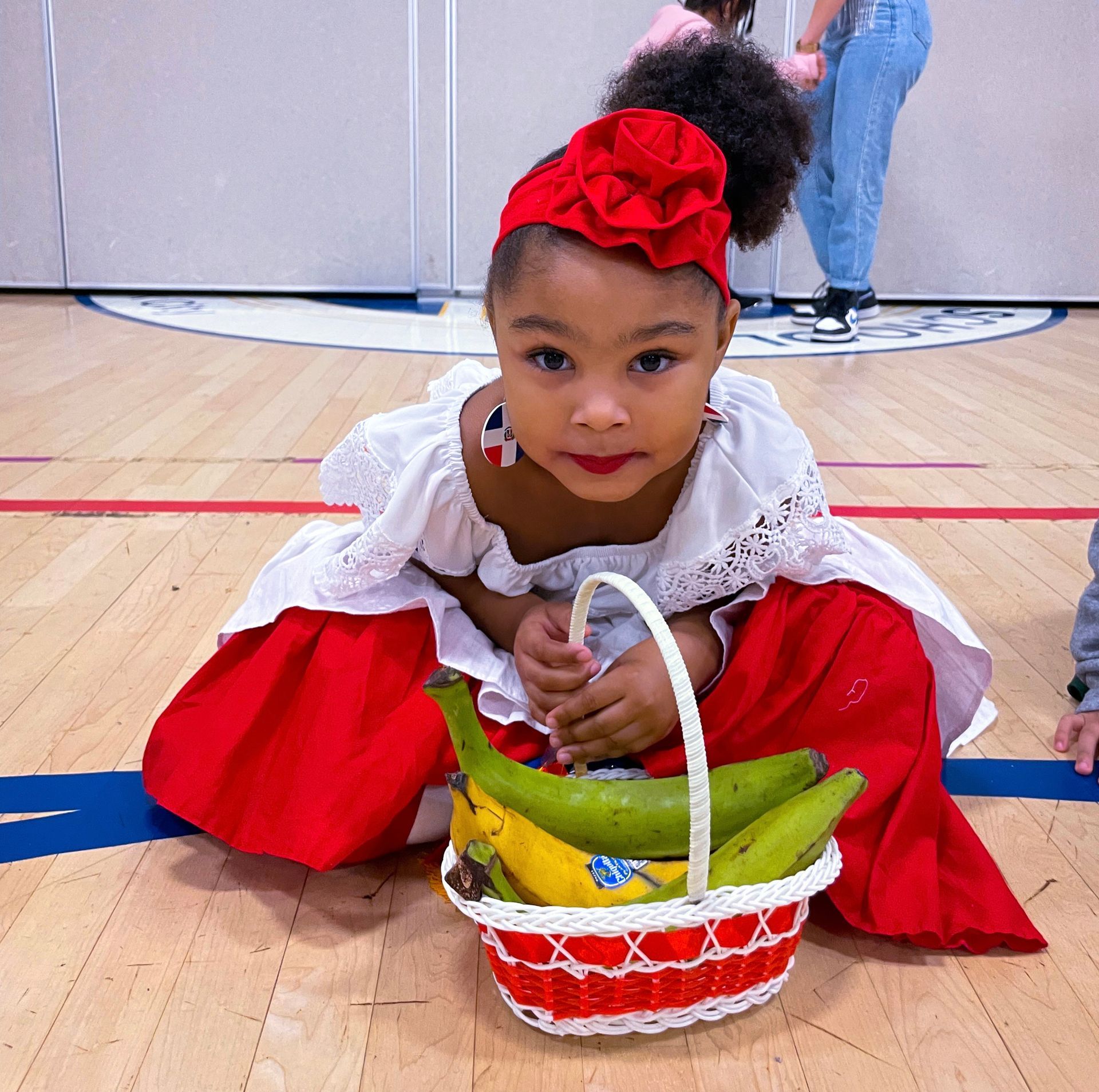 Child in a red dress kneels by a basket of Easter eggs on a gym floor, holding a white ribbon.