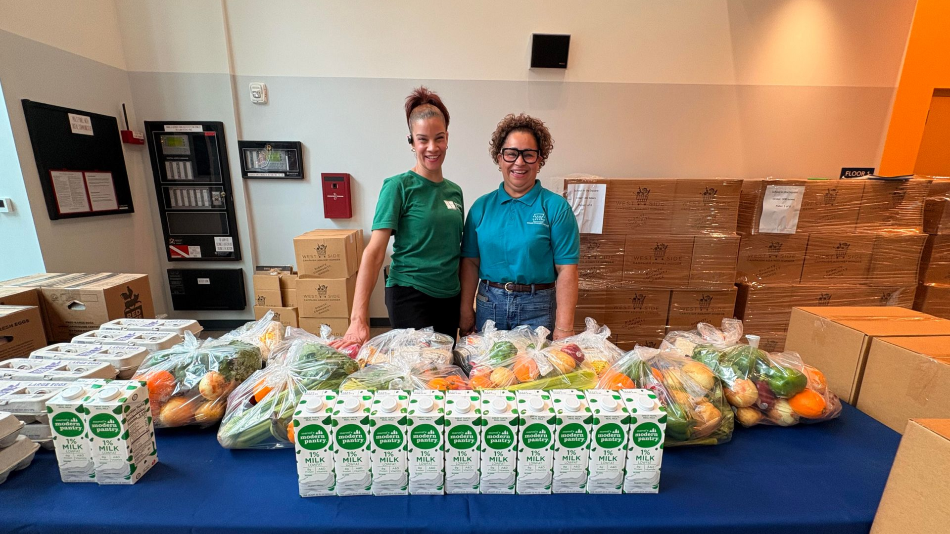 Two people stand behind a table of produce and boxed goods in a lobby or hall.