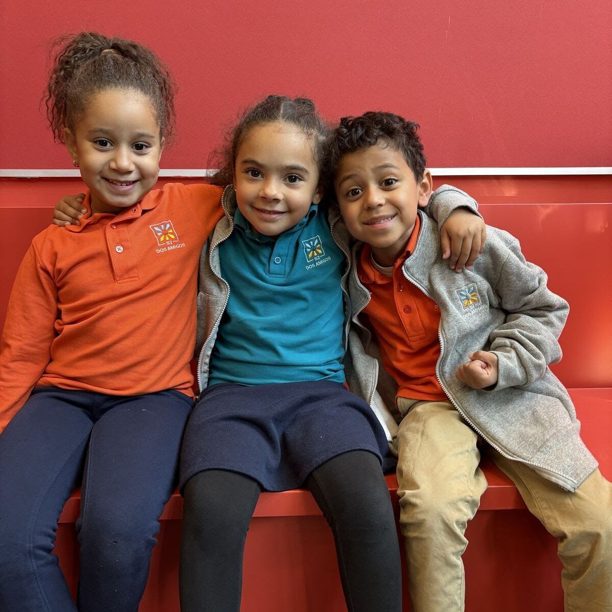 Three children sit together on a red bench, smiling, with arms around each other.