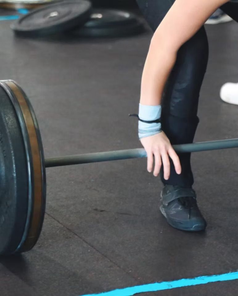 Person in black pants reaching toward a barbell on a gym floor, with blue wrist wraps.