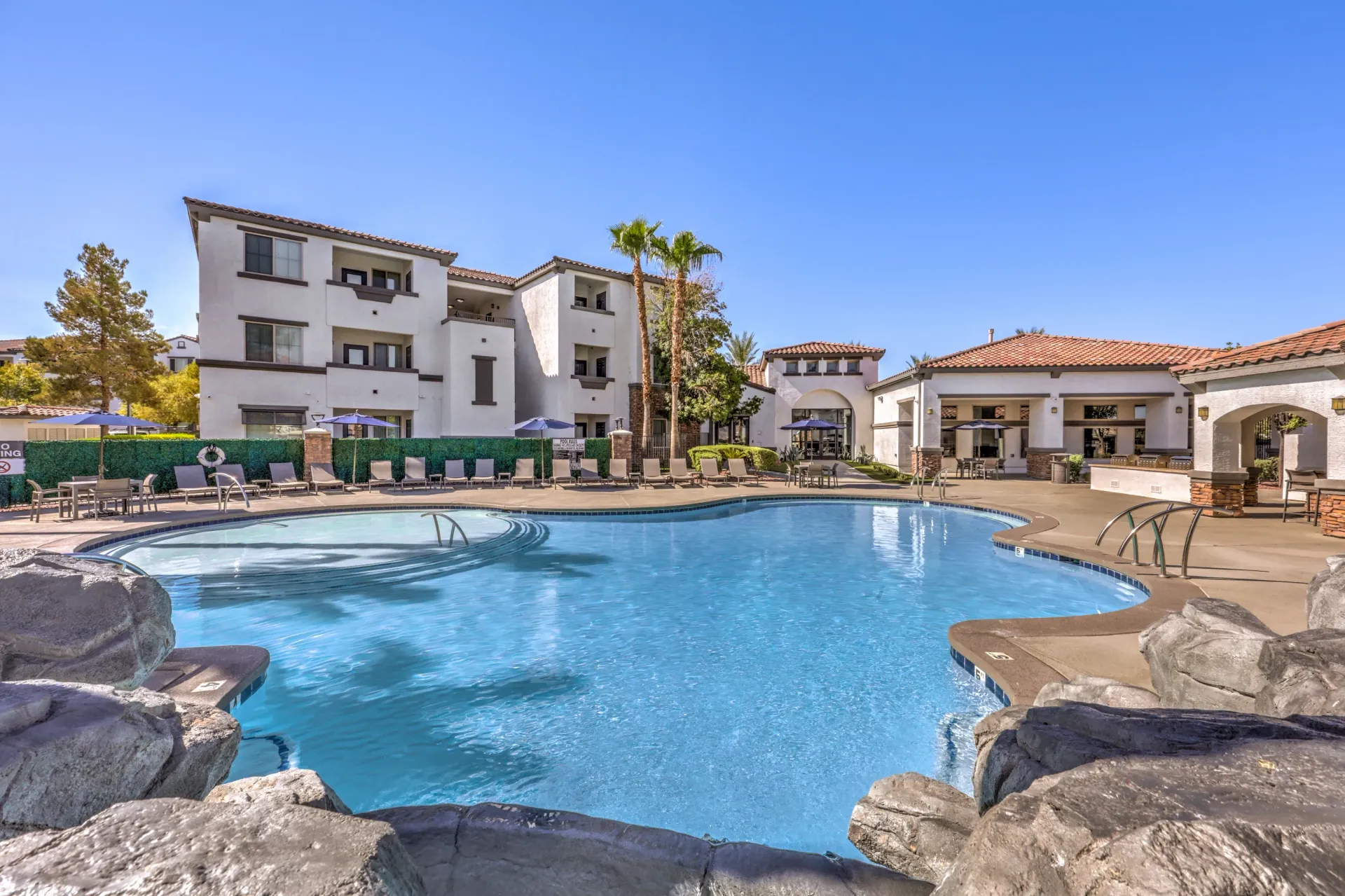 Resort-style swimming pool with lounge chairs and umbrellas, next to apartment buildings at Cornerstone Park in Henderson, NV.