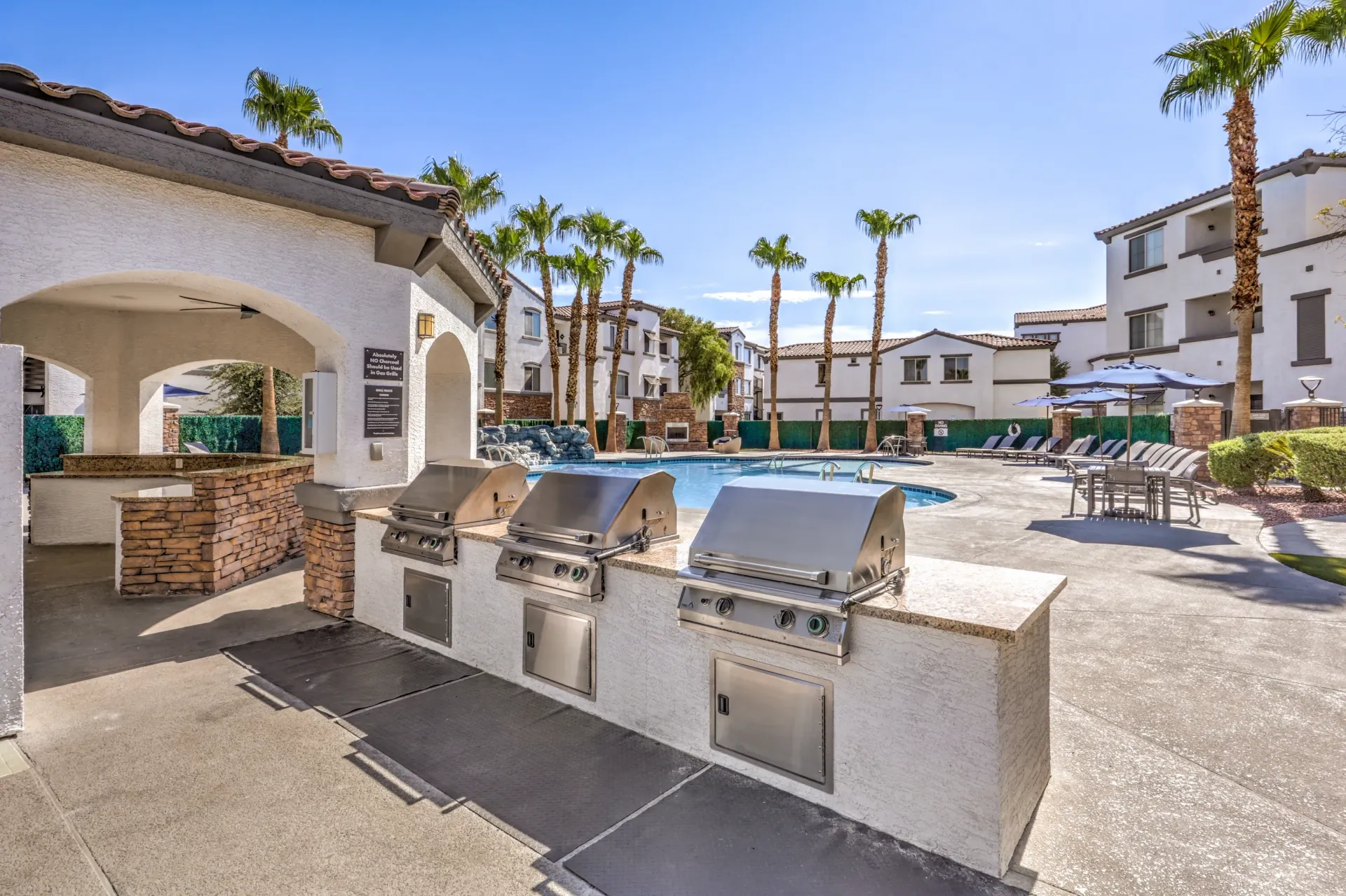 Outdoor grills and swimming pool area with lounge chairs and palm trees at Cornerstone Park in Henderson, NV.