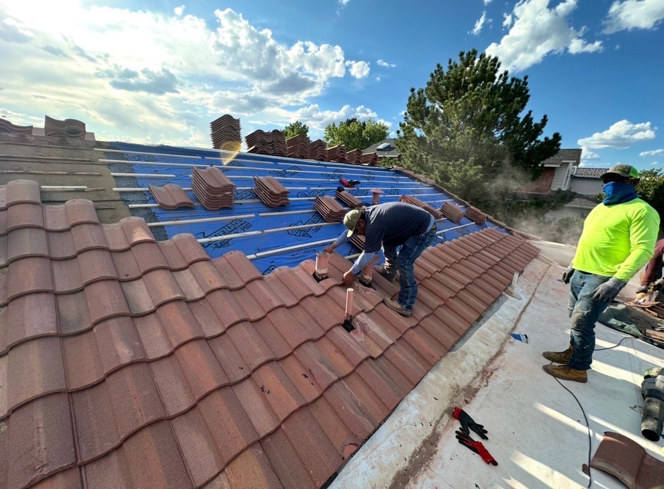 Two roofers installing red clay tiles on a rooftop under a partly cloudy sky. One wears a face covering.
