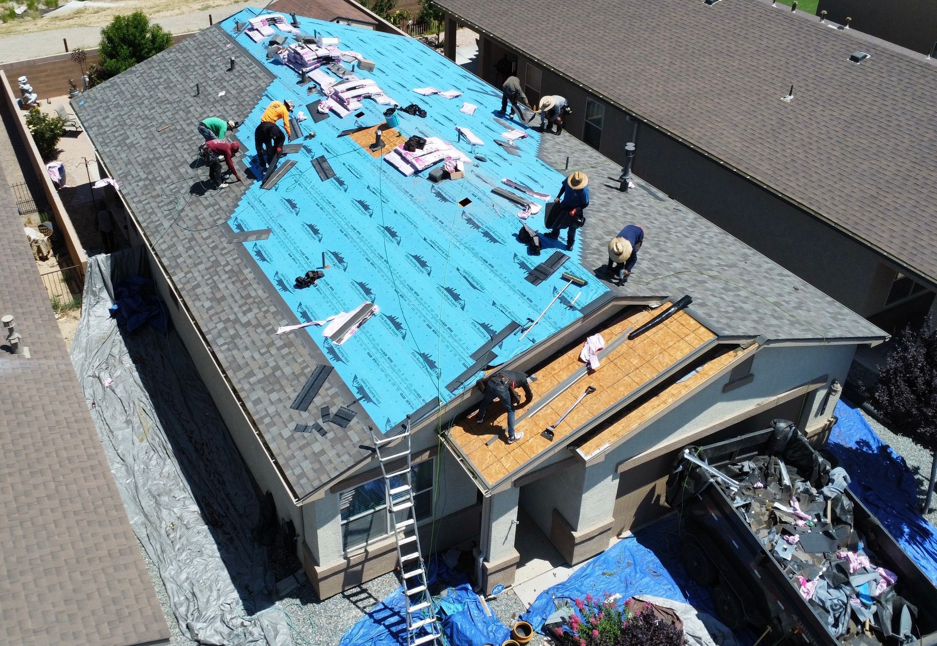 Roofers replacing shingles on a house; blue underlayment visible, dumpsters beside the home.