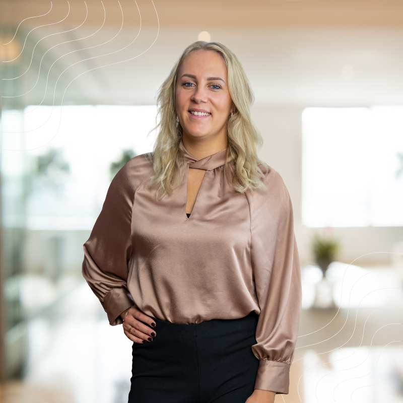 Vrouw met blond haar, glimlachend, gekleed in een roségouden blouse en een zwarte broek, in een lichte kantooromgeving.