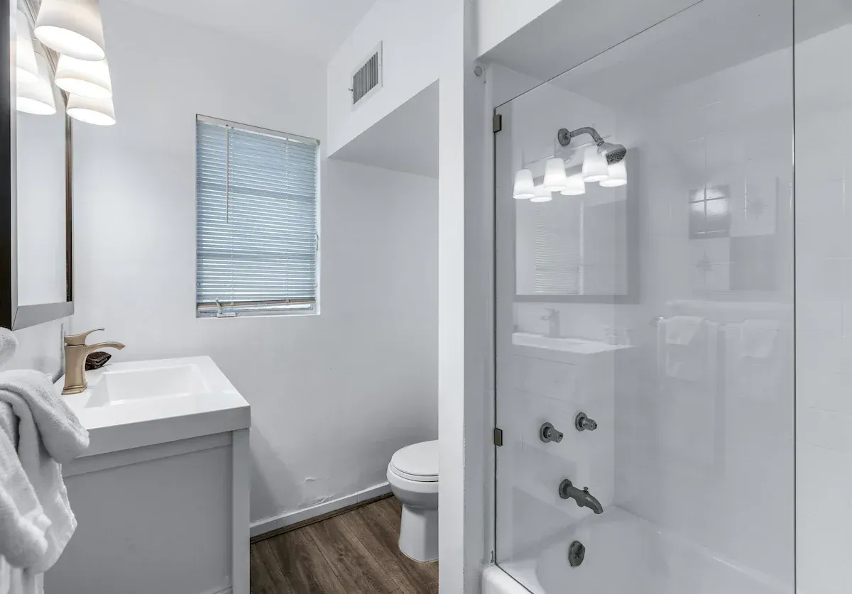 Bright modern bathroom with white vanity, toilet, and glass shower, accented by wood flooring and a small window.