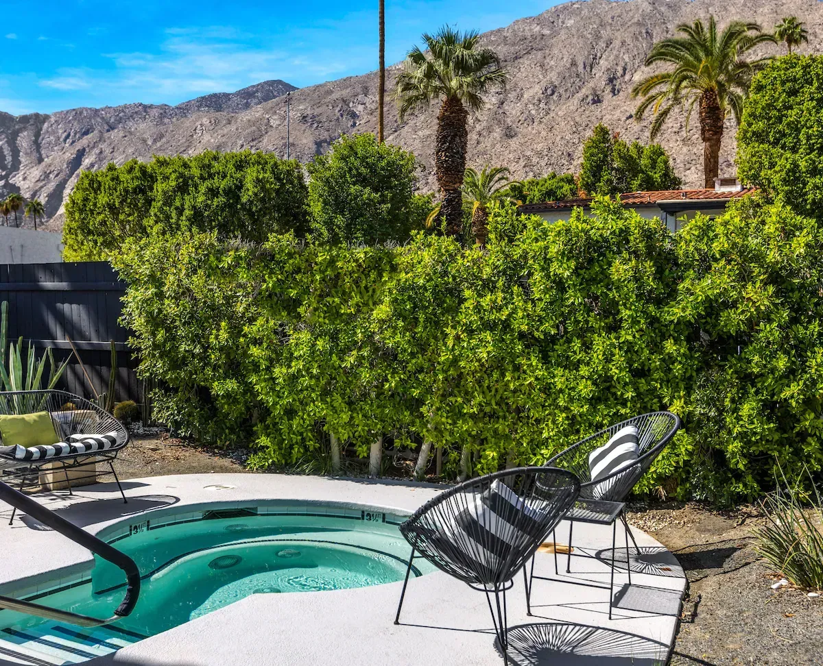 Poolside patio with turquoise spa, black lounge chairs, tall hedge, and desert mountains in the background