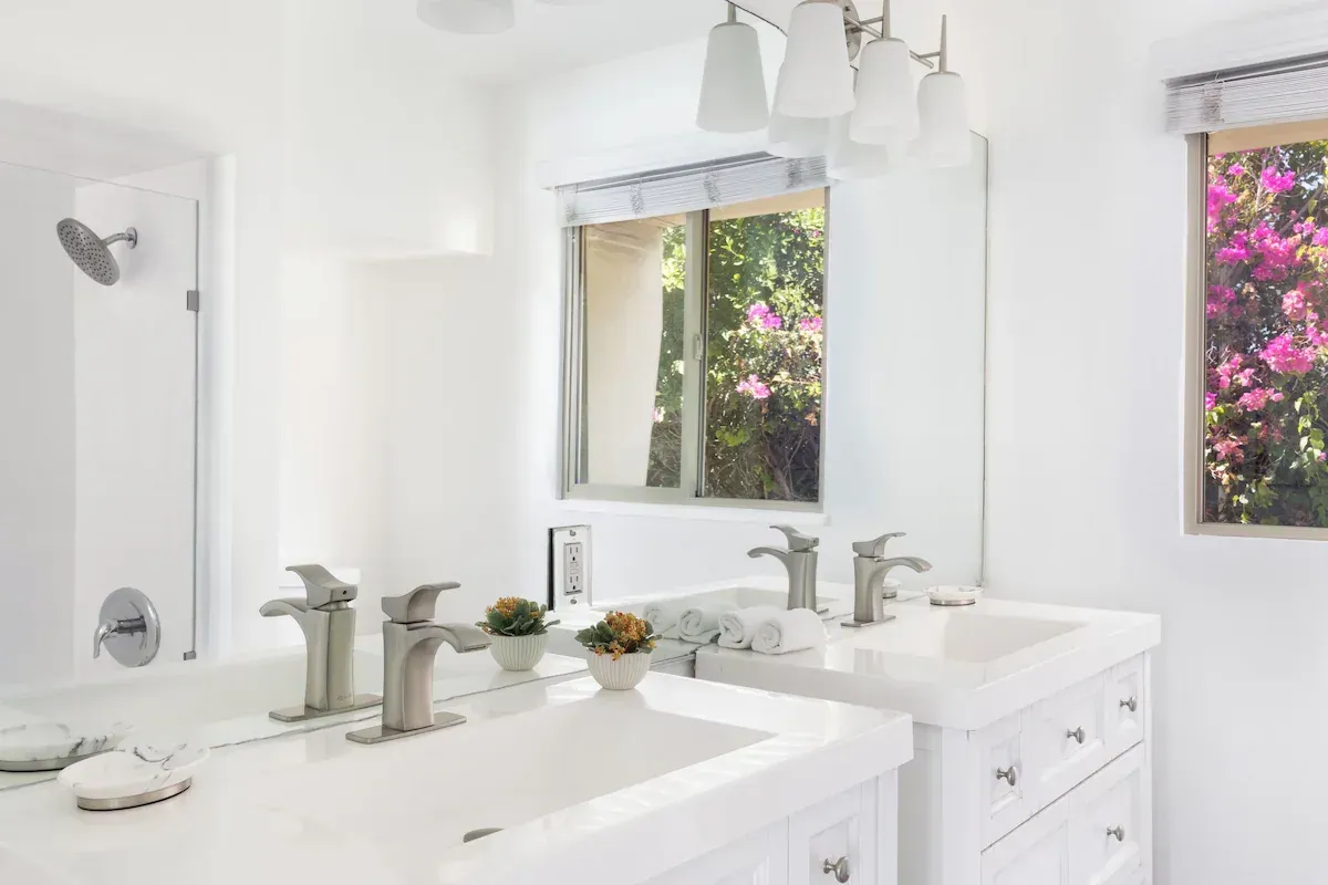 Bright white bathroom with double sinks, mirrors, and floral views through windows.