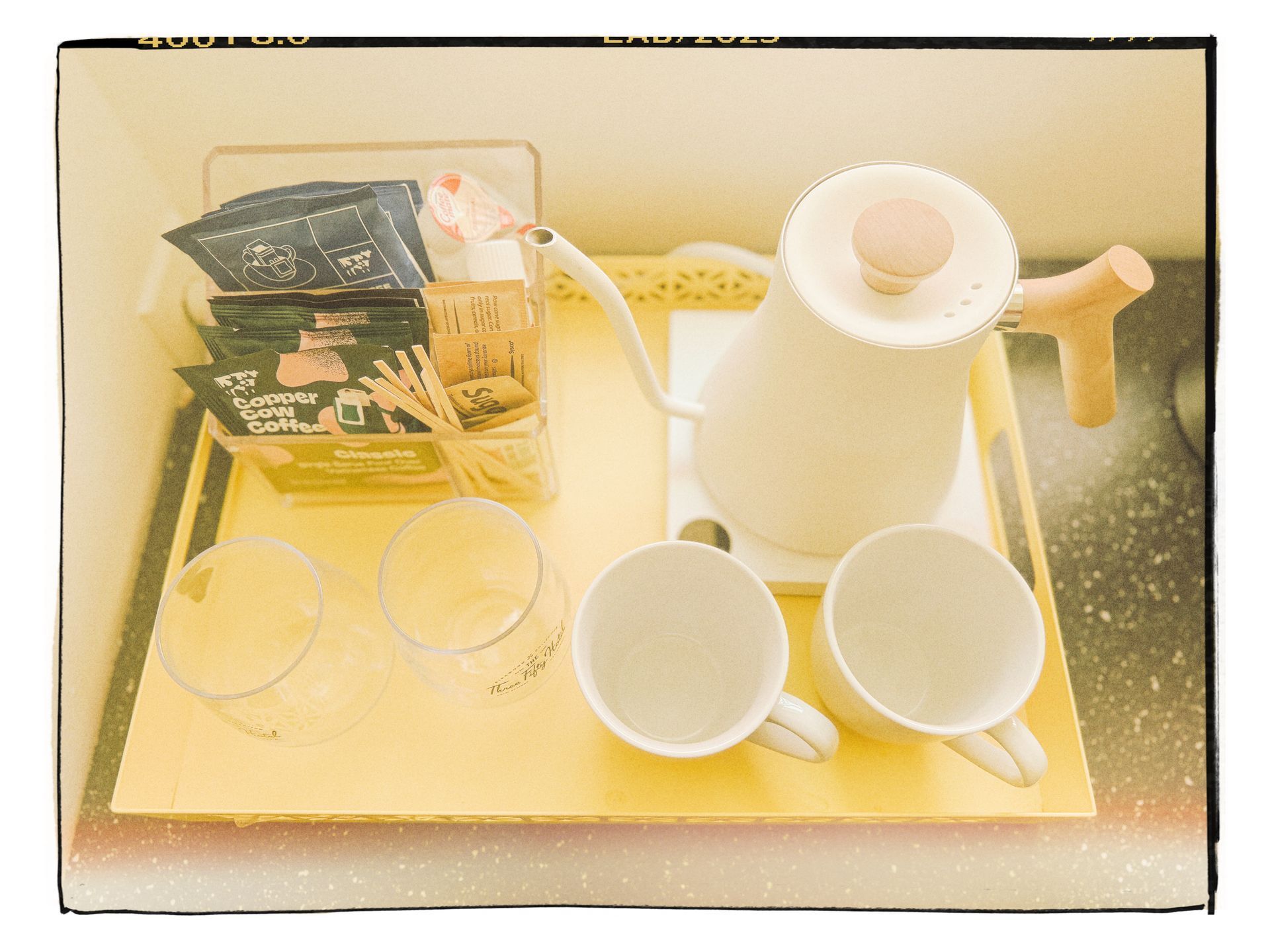 Tray with a white teapot, two cups, glasses, and snack packets on a table