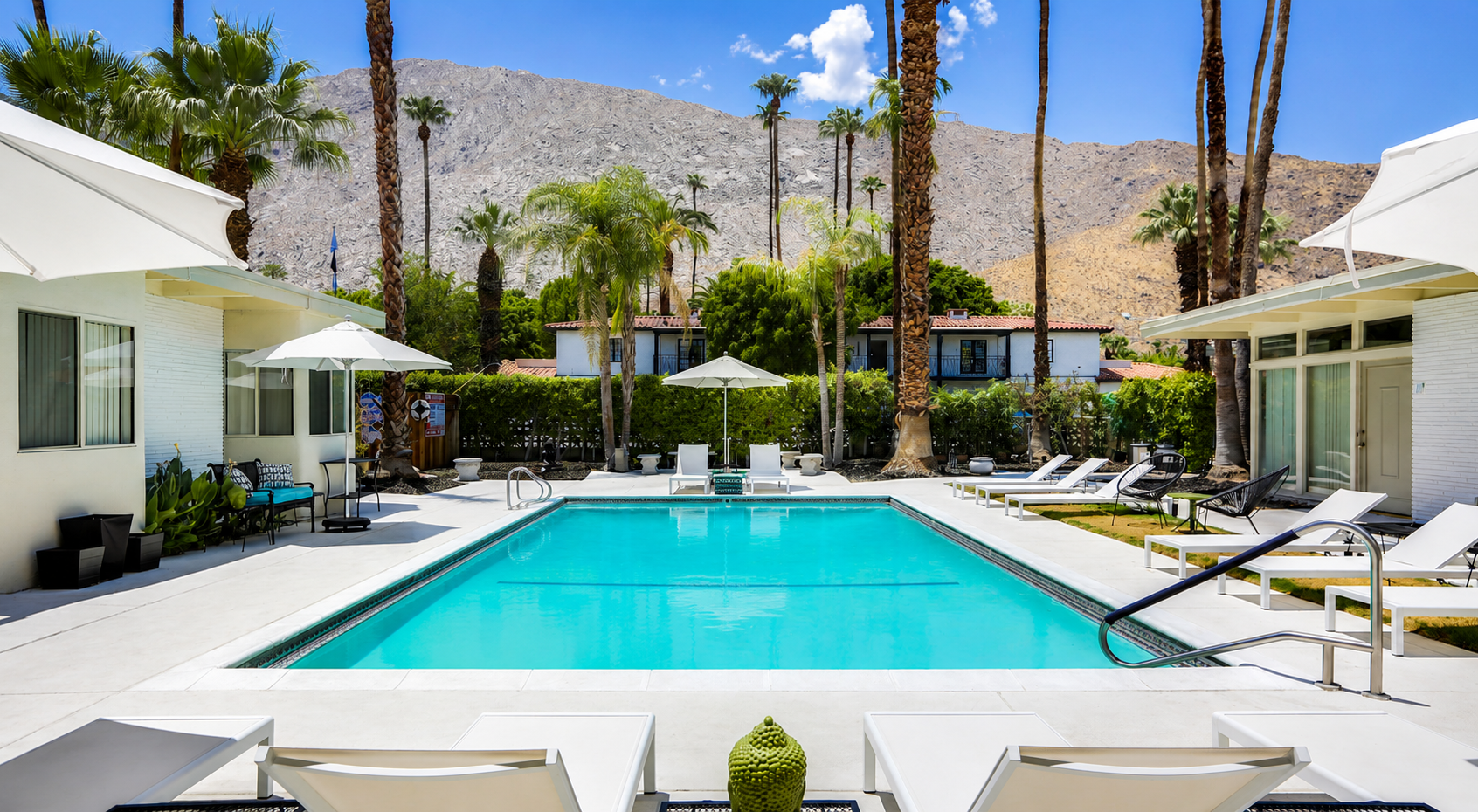 Modern resort pool courtyard with white loungers, palm trees, and desert mountains in the background