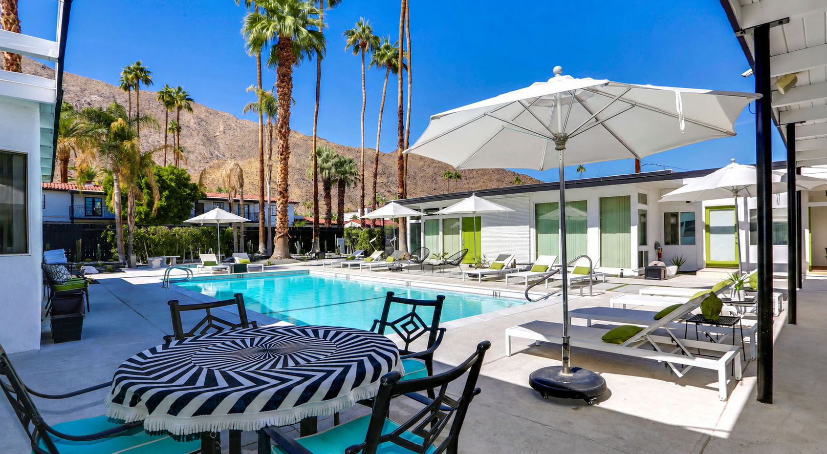 Sunlit resort patio with striped table, lounge chairs, umbrella, pool, and palm trees against mountains
