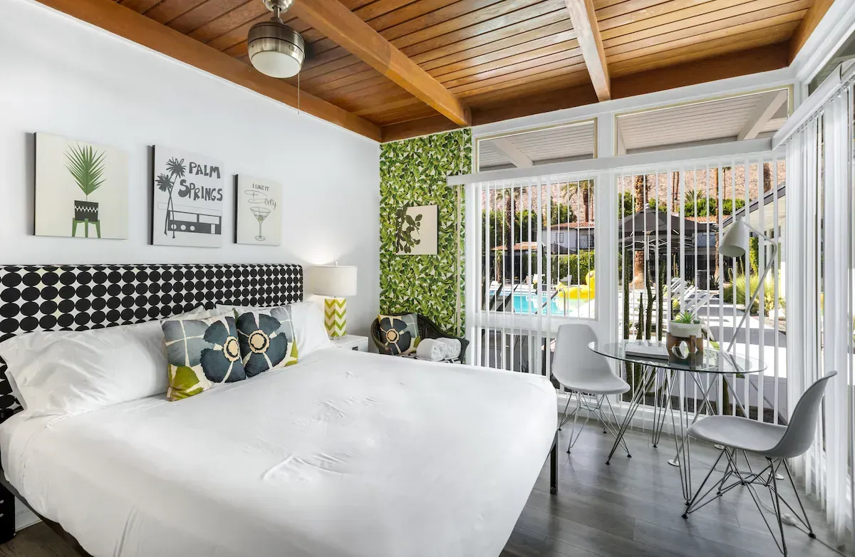Bright bedroom with white bed, patterned pillows, and a sunlit balcony view through glass doors