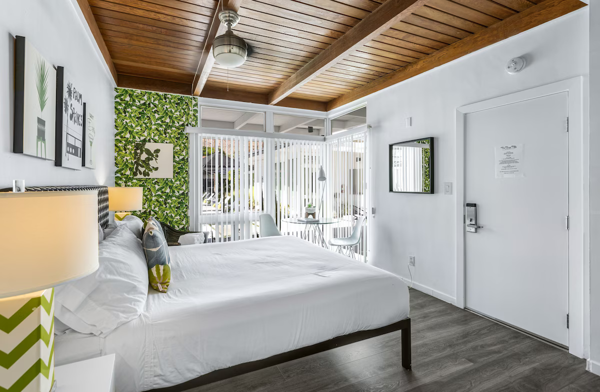 Bright bedroom with white bedding, wood ceiling, leafy wall art, and sliding glass doors to a balcony.