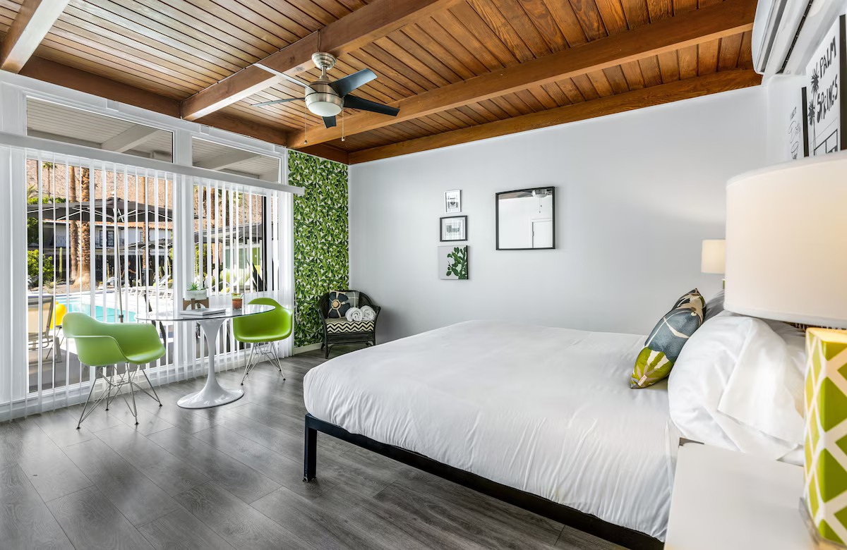 Modern bedroom with white bed, wood ceiling, and sliding doors opening to a patio with green chairs