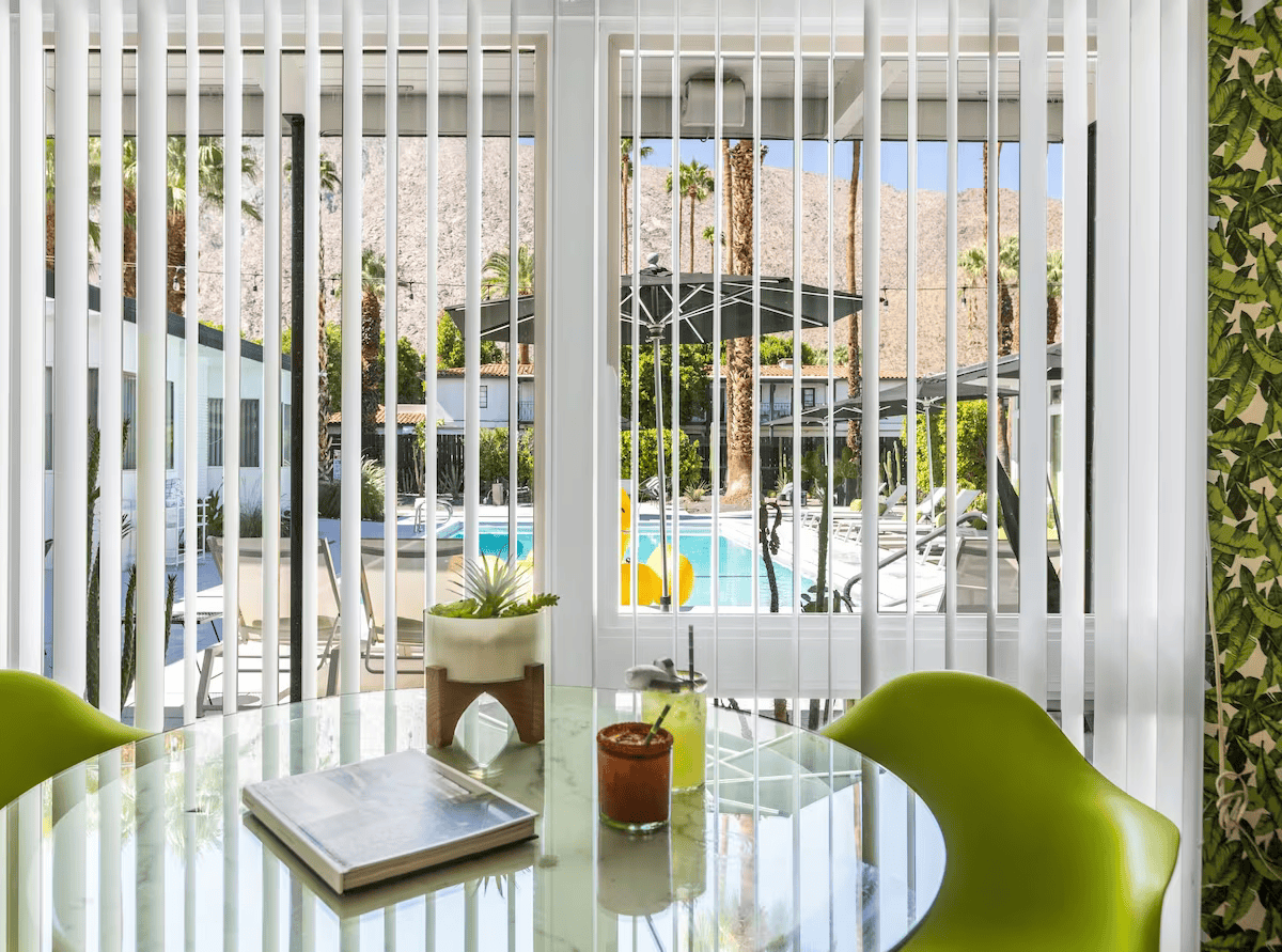 Bright patio with a glass table, lime-green chairs, and white vertical blinds overlooking a sunny pool area