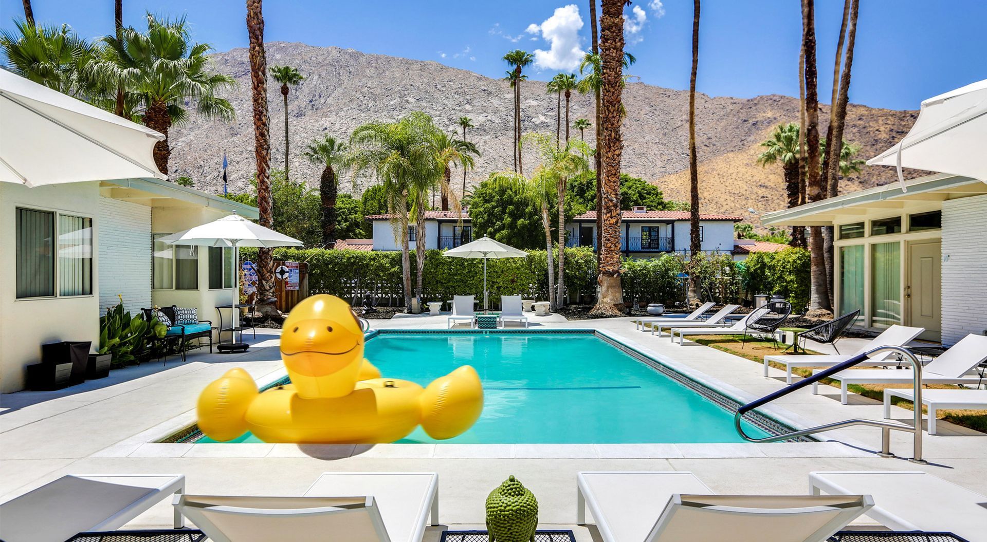 Resort pool with large yellow duck float, palm trees, lounge chairs, and desert mountains in the background