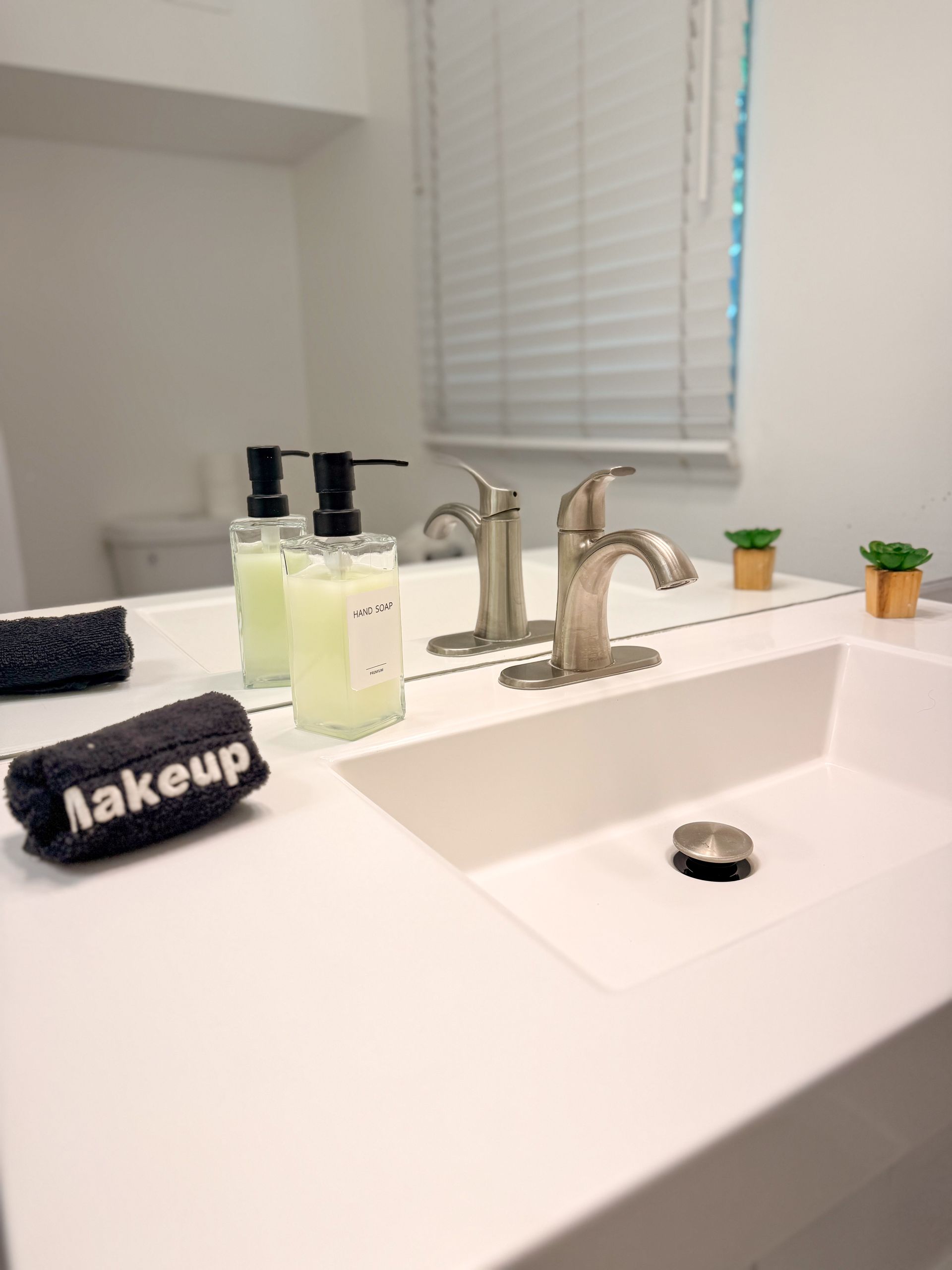 Bathroom vanity with sink, soap dispensers, makeup bag, and small potted plants by a mirror