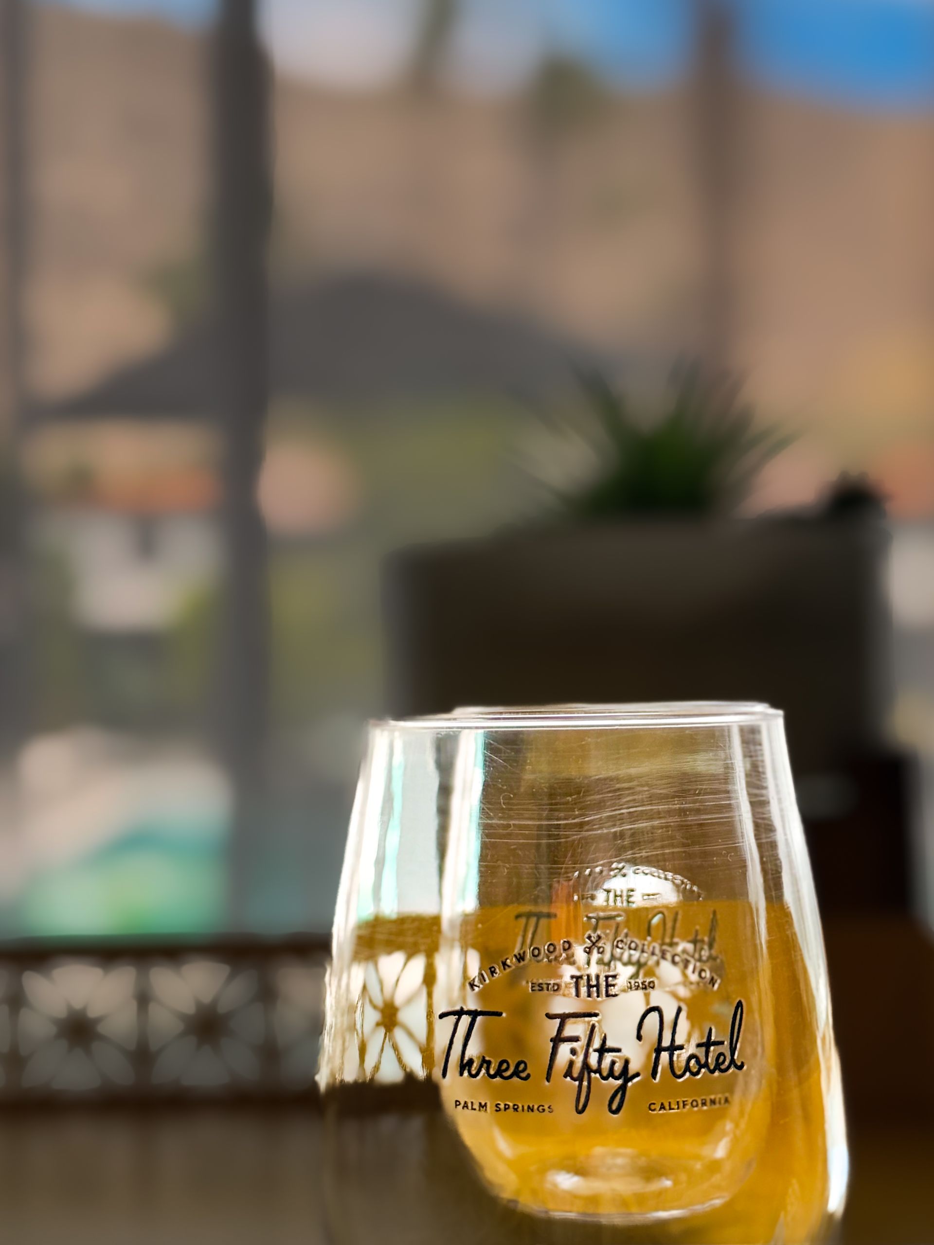 Stemmed glass of amber drink on a table, with a blurred window and plant in the background