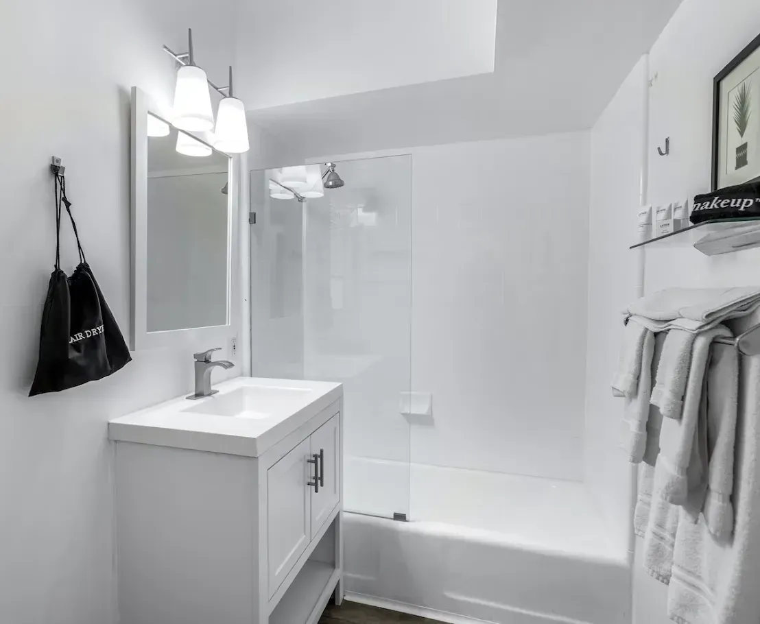 Bright white bathroom with vanity, mirror, walk-in shower, and towels hanging on the wall.