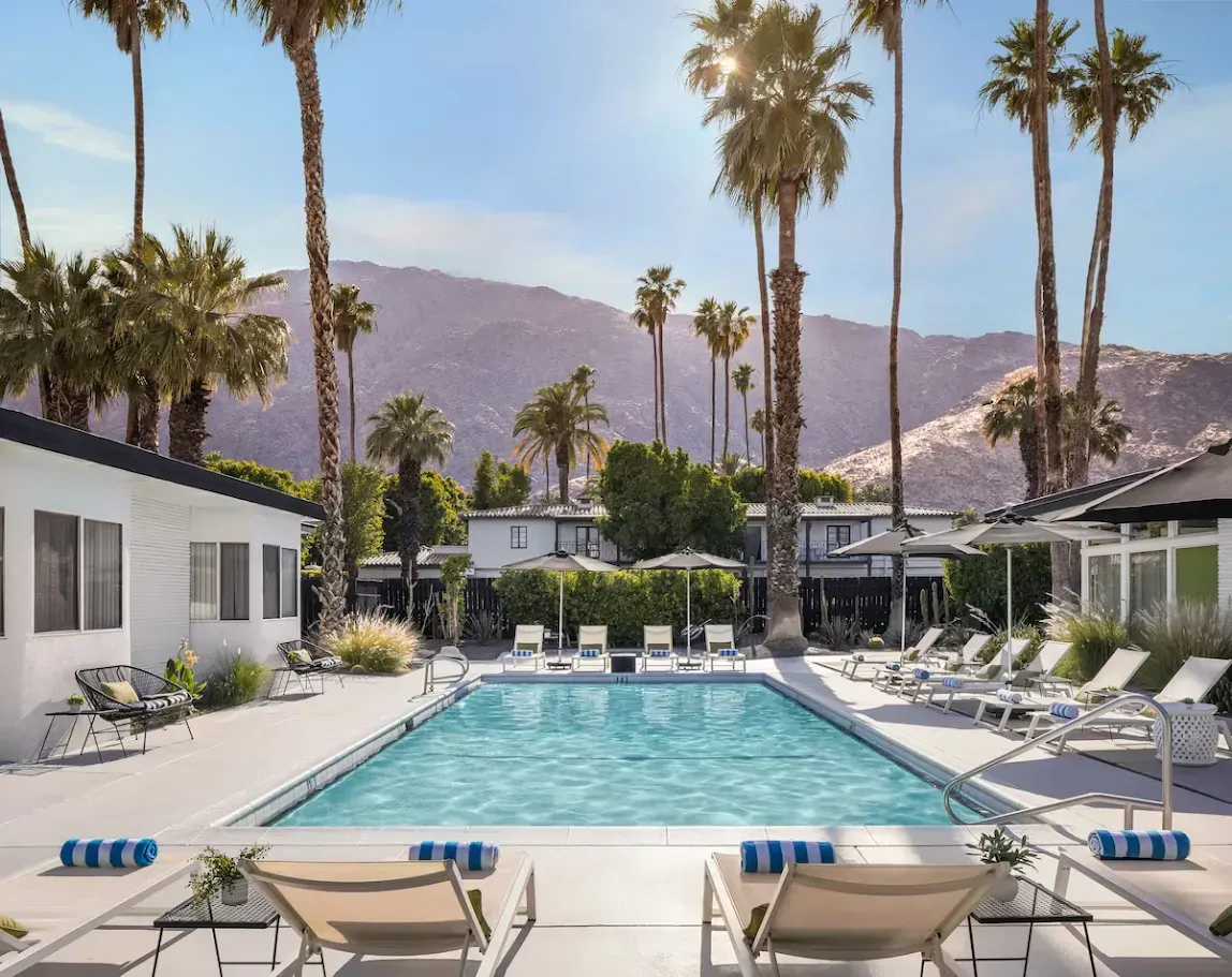 Modern resort pool with lounge chairs, white buildings, palm trees, and mountains in the background