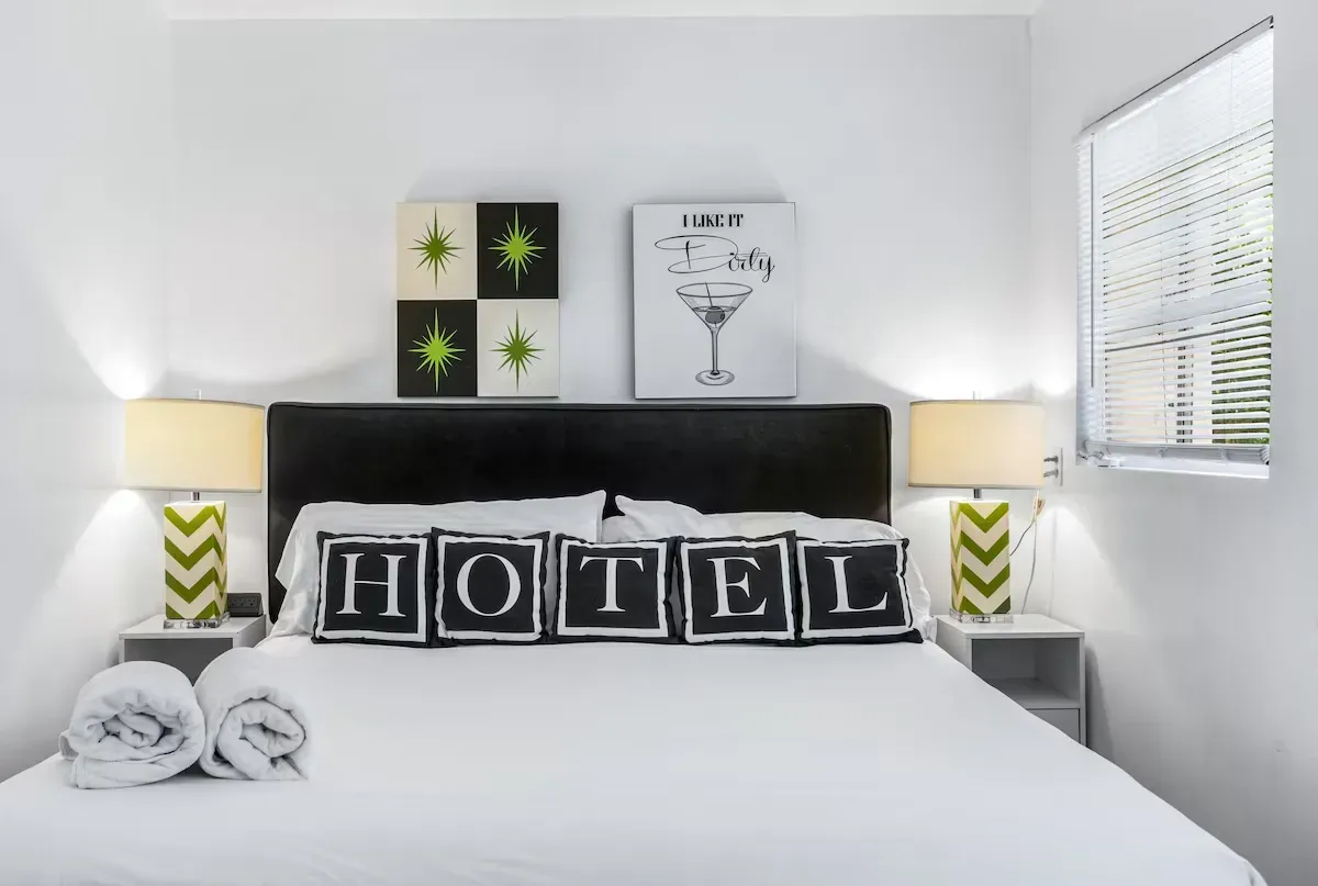 Bright hotel-style bedroom with a bed, black “HOTEL” pillows, and matching bedside lamps