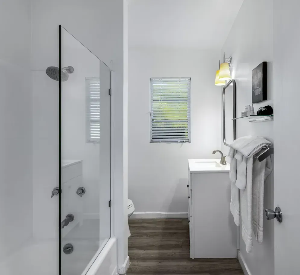 Narrow white bathroom with glass shower, vanity sink, towel rack, and small window at the end