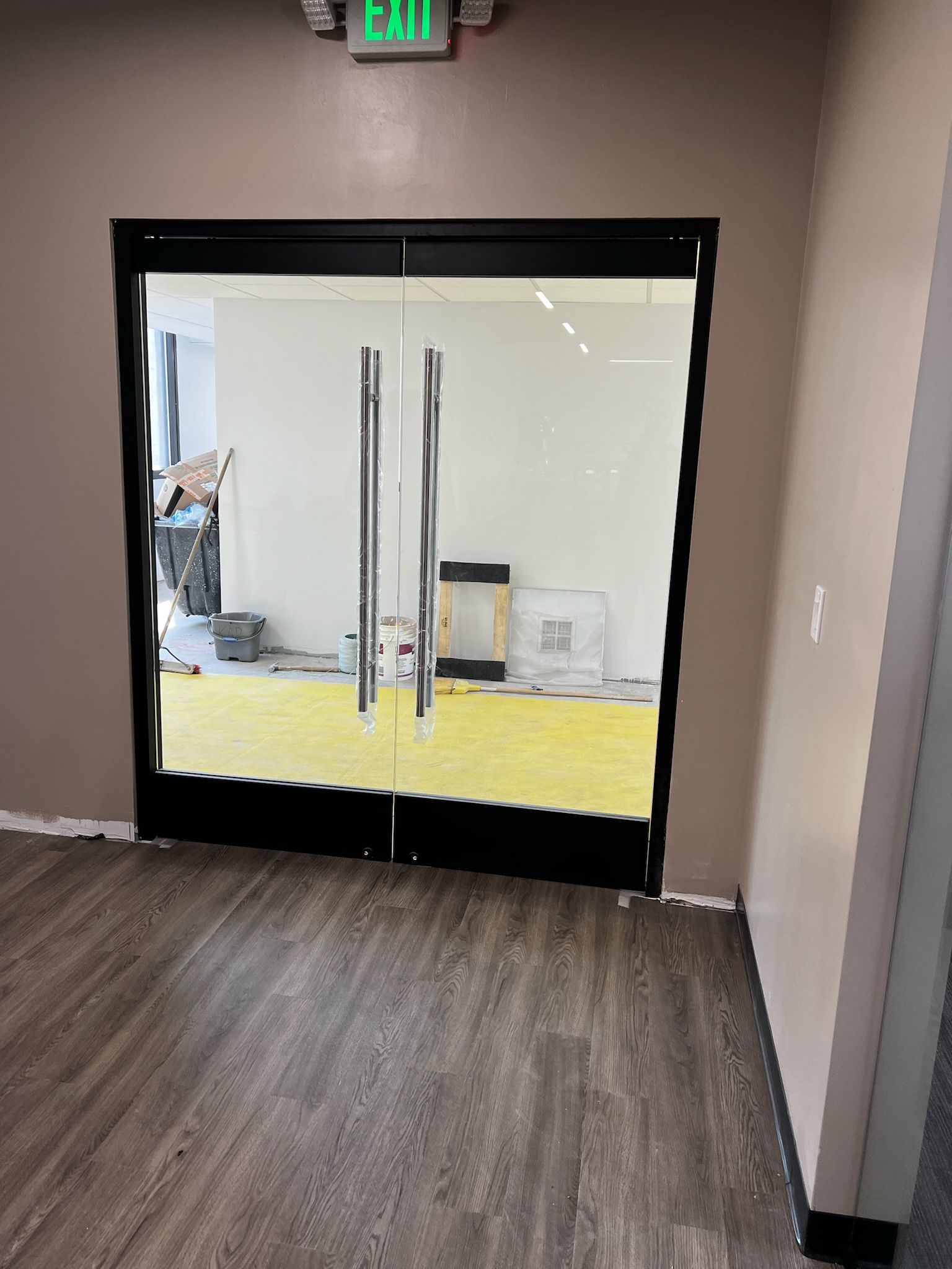 Glass double doors with chrome handles, black frame, and a beige wall. Exit sign above.