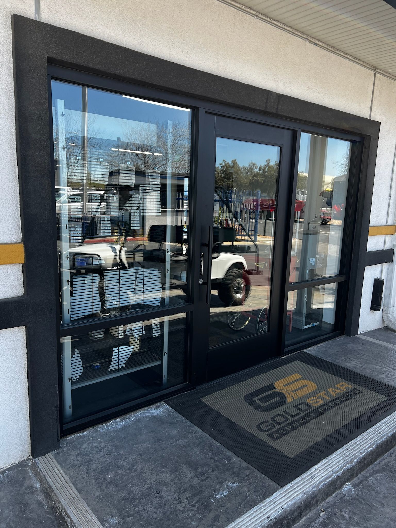 Black framed glass doors with gold doormat, leading into a building.