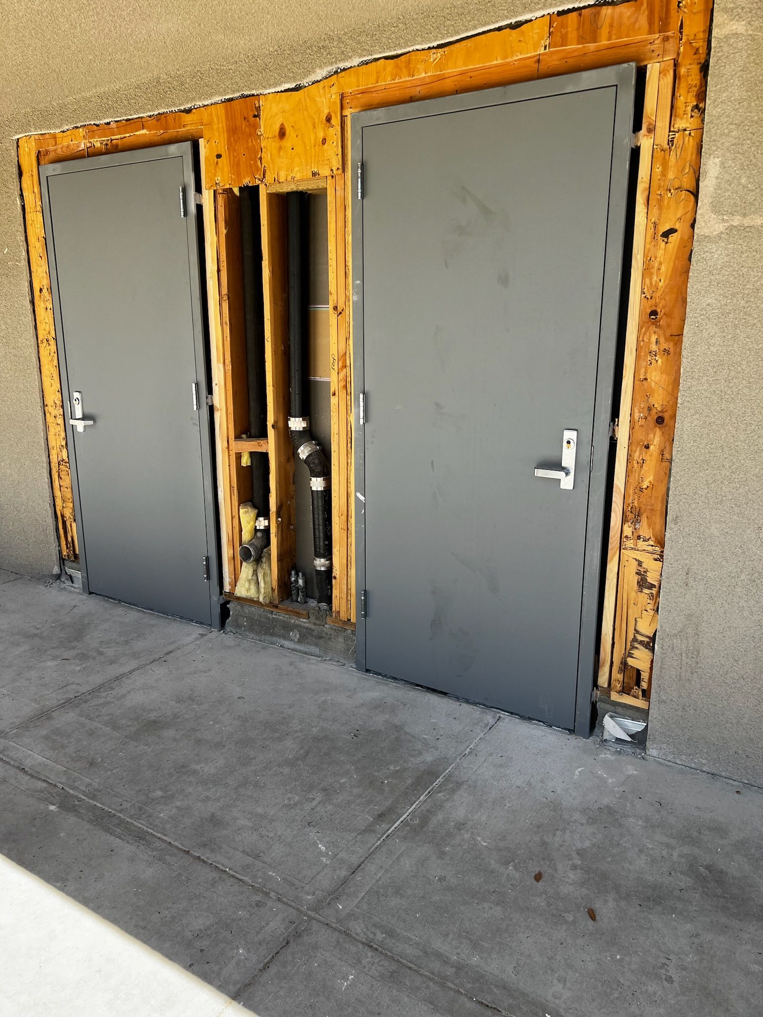Two grey doors set in a damaged wooden frame, next to a concrete ground, against a tan wall.