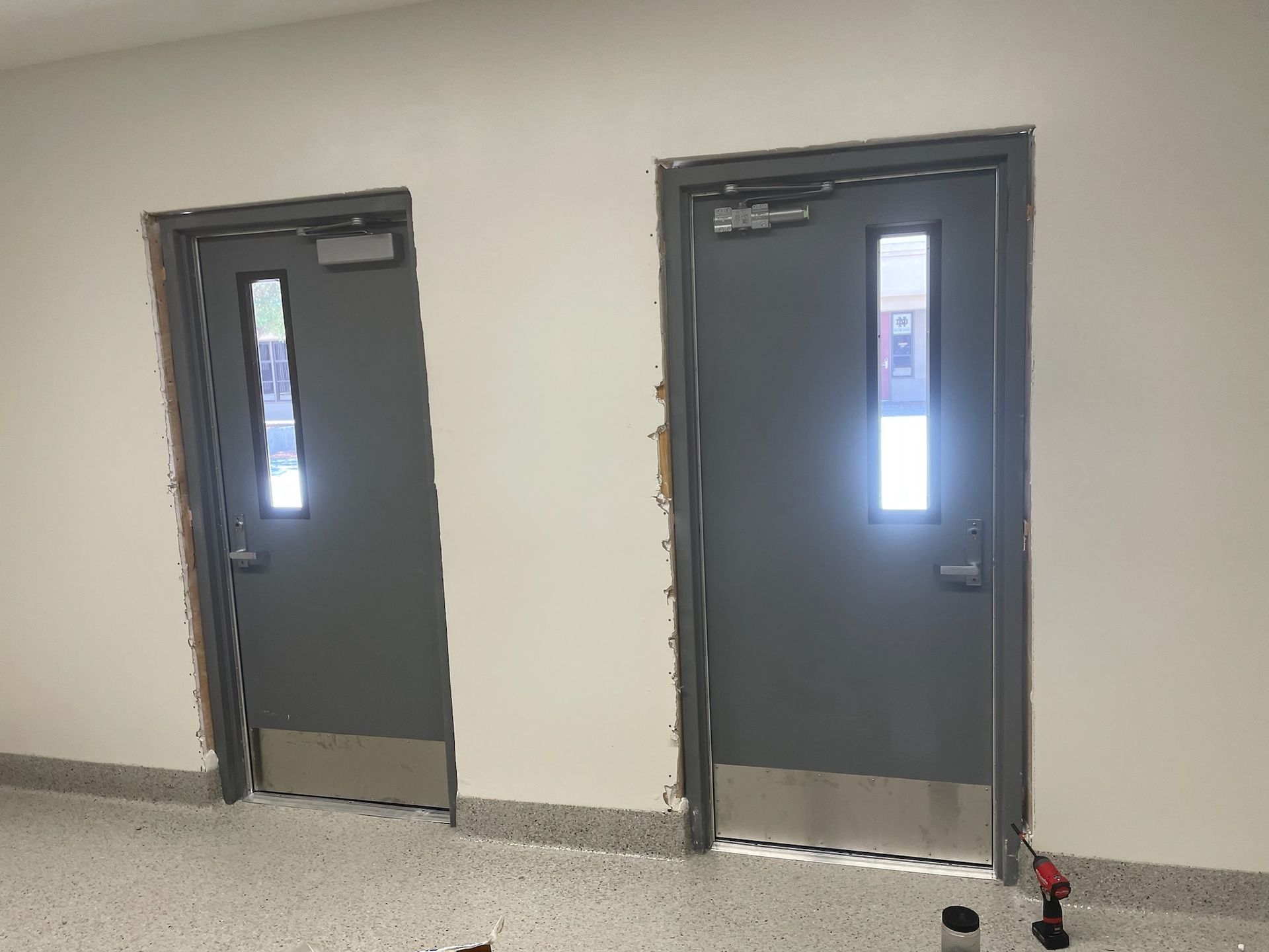 Two gray doors with windows in a white hallway, stainless steel kick plates, and hardware.