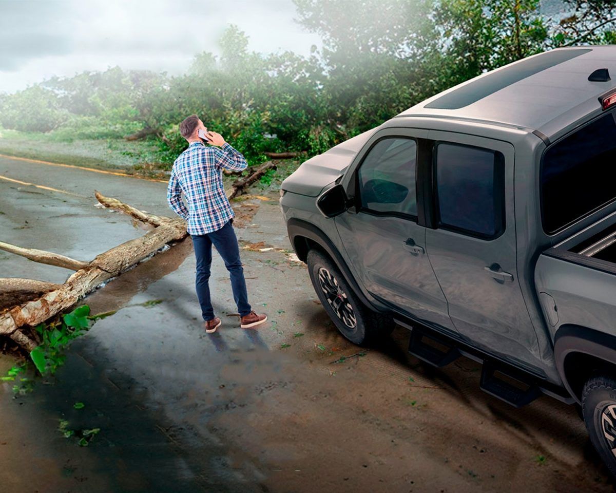 Persona junto a una camioneta gris frente a un árbol caído que bloquea una carretera inundada.