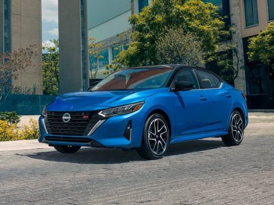 Un sedán Nissan azul estacionado al aire libre cerca de edificios modernos y árboles.