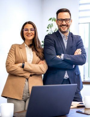 Dos compañeros sonrientes, de pie detrás de un ordenador portátil en una oficina luminosa, con los brazos cruzados.