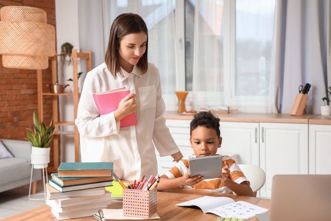 A woman is standing next to a boy sitting at a table using a tablet.