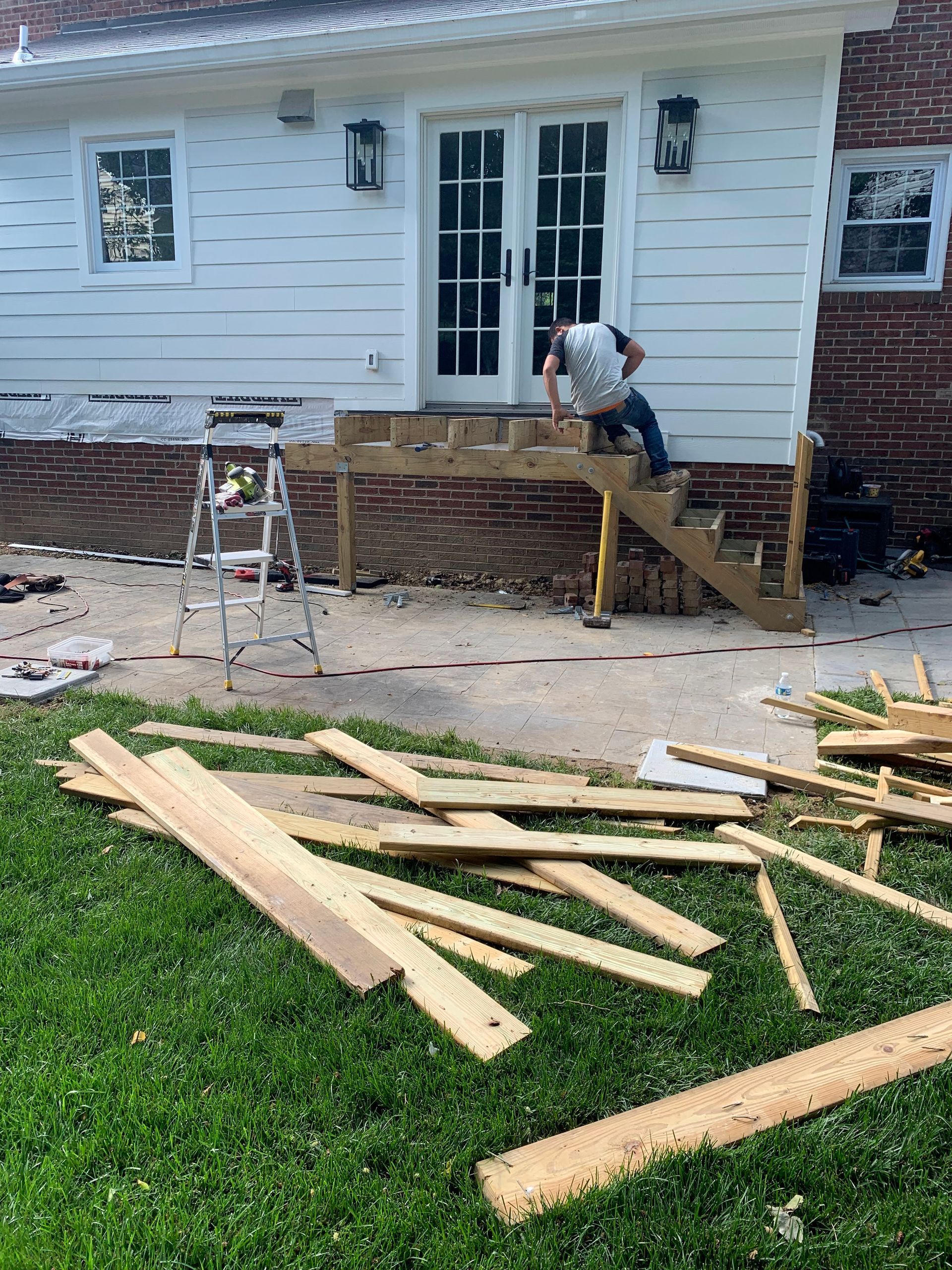Person building wooden steps outside a white house with a brick foundation; boards on the grass.
