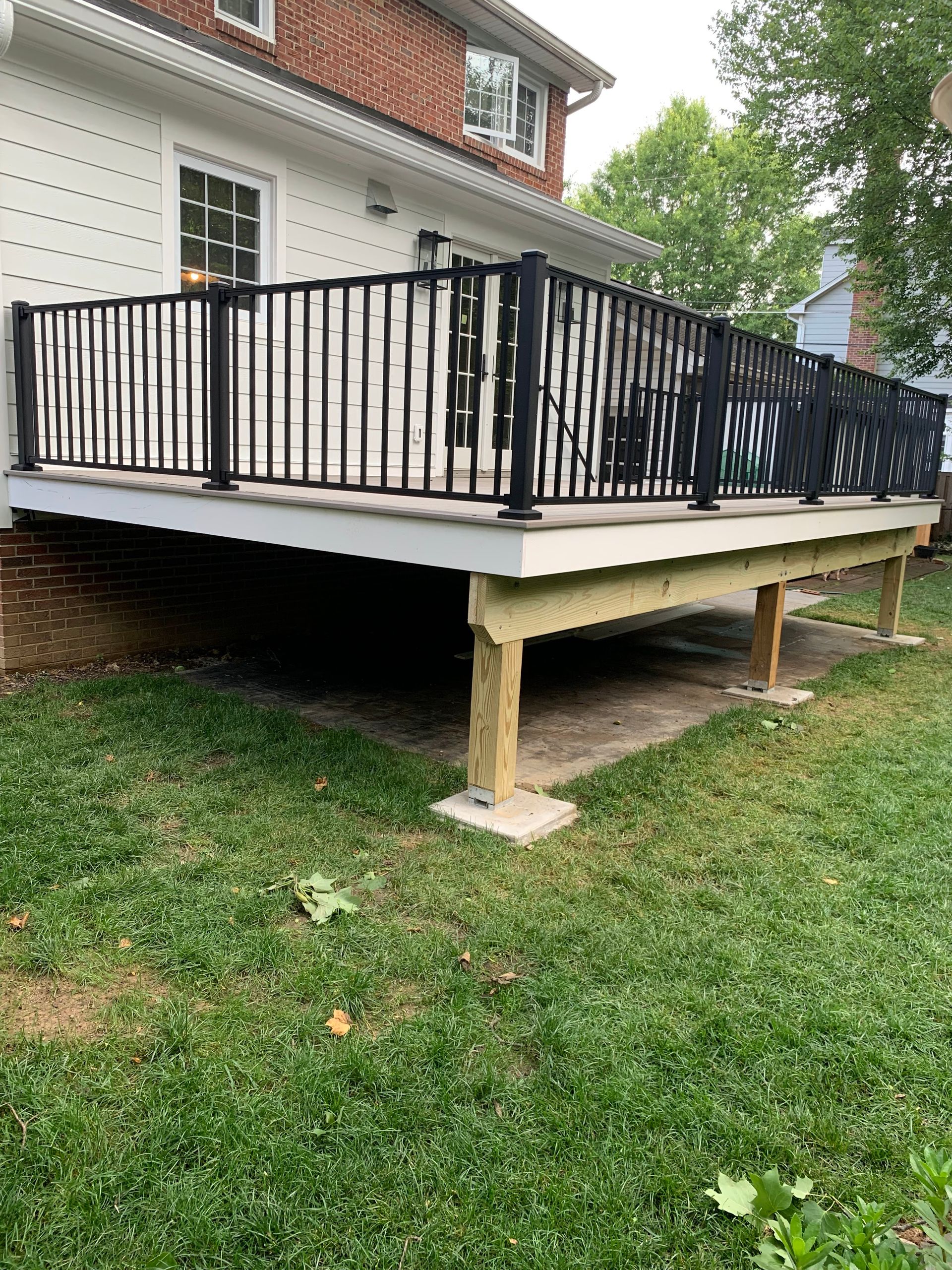 A deck with black railings and a white base, supported by wooden posts on concrete blocks, set in a grassy yard.