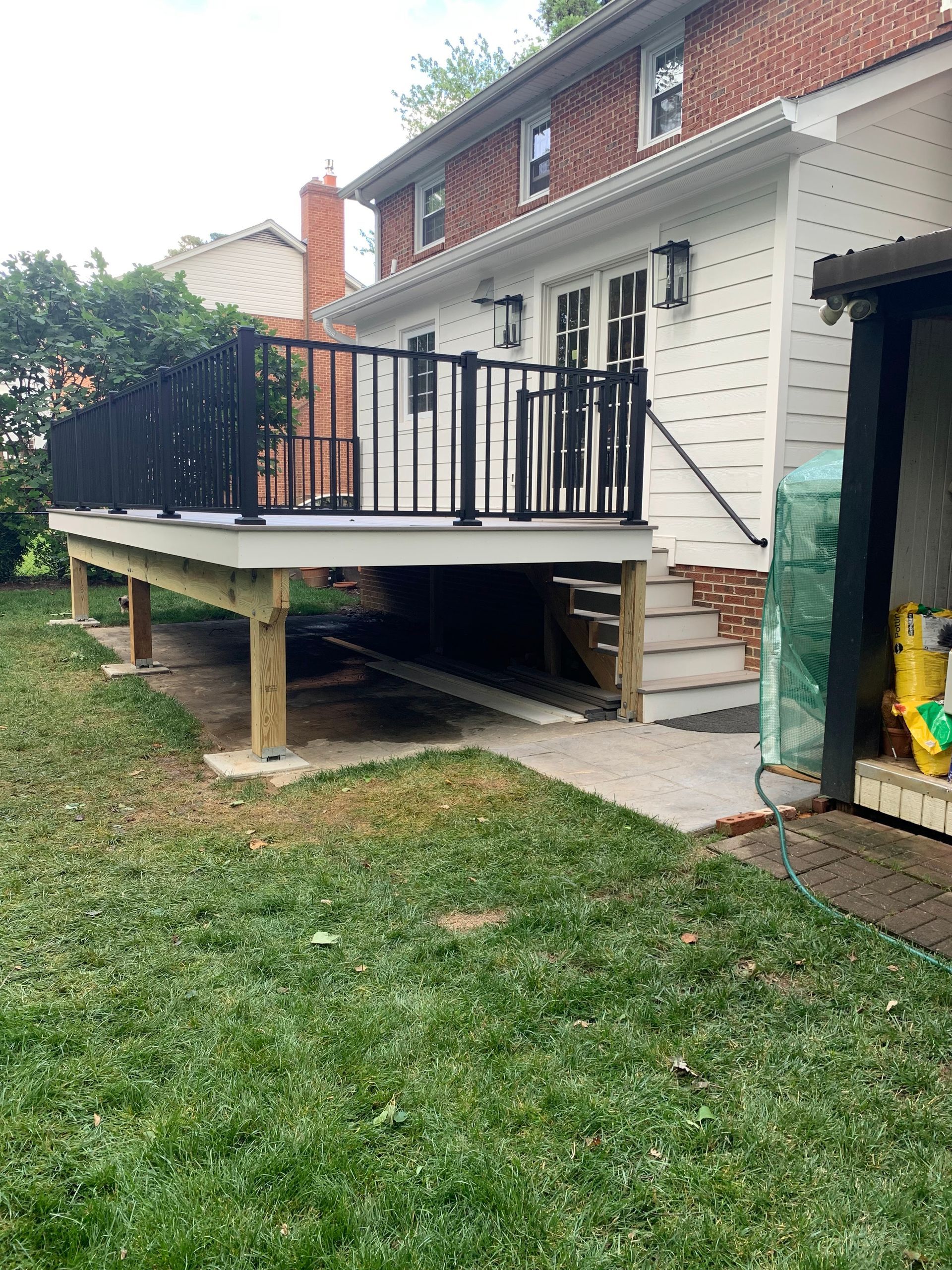 Backyard deck attached to a brick house. Black railing, grey deck surface. Underneath are wooden supports on concrete blocks.