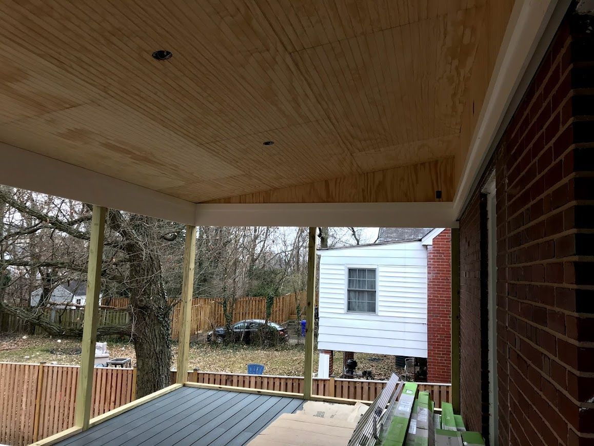 Newly constructed porch with wood ceiling and brick wall next to it; view of neighborhood in the background.