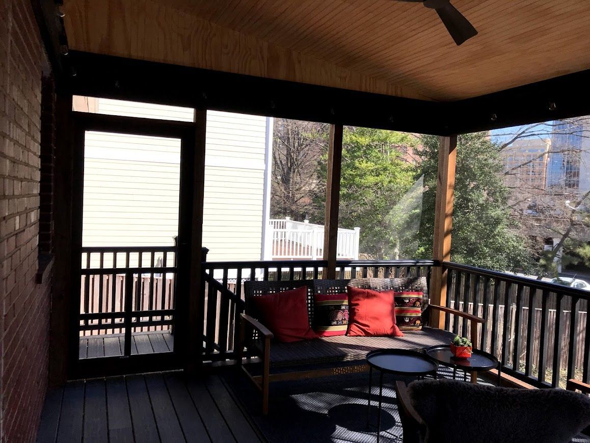 Screened porch with dark wood trim, furniture, red pillows, and a view of trees and a building.