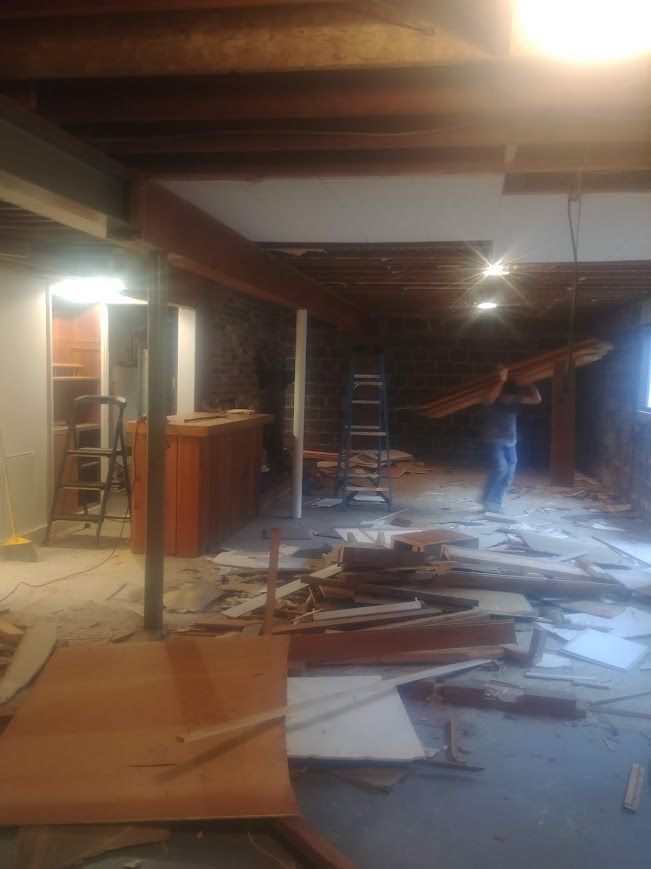 Basement renovation in progress; debris on floor, person carrying wood, exposed beams and support columns.