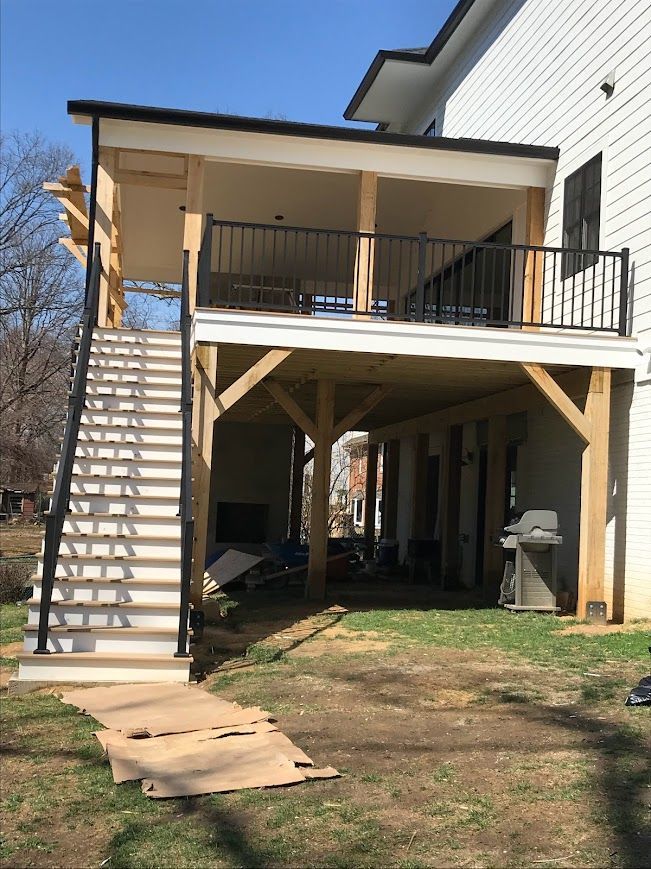 Two-story outdoor deck with stairs; black railing, wooden supports, and white siding. Backyard setting.
